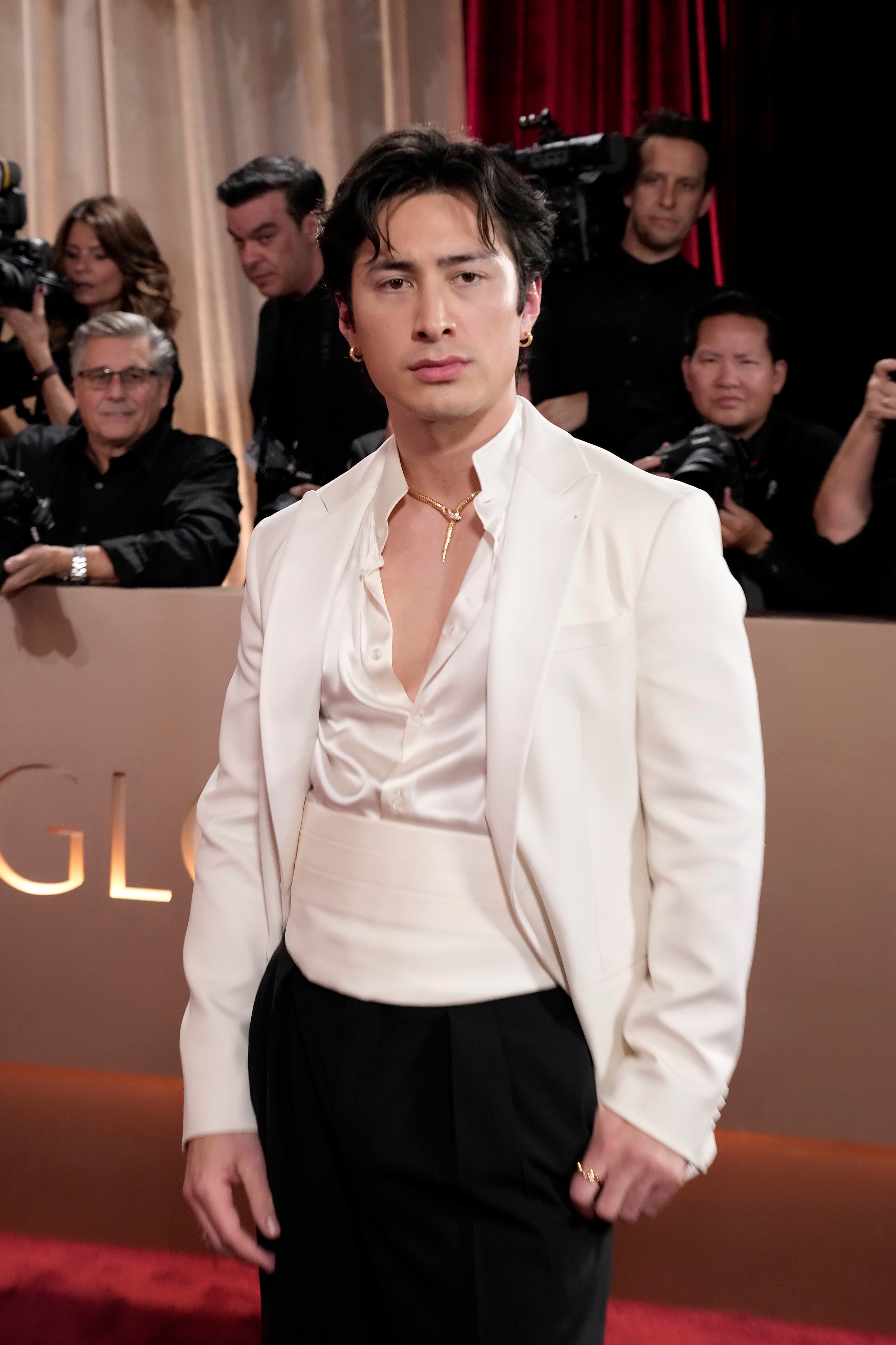 Person på rød løper iført en stilig kremdress med dyp skjorte med V-hals, poserer for bilder. Bakgrunnen viser fotografer og et bakteppe for hendelsen