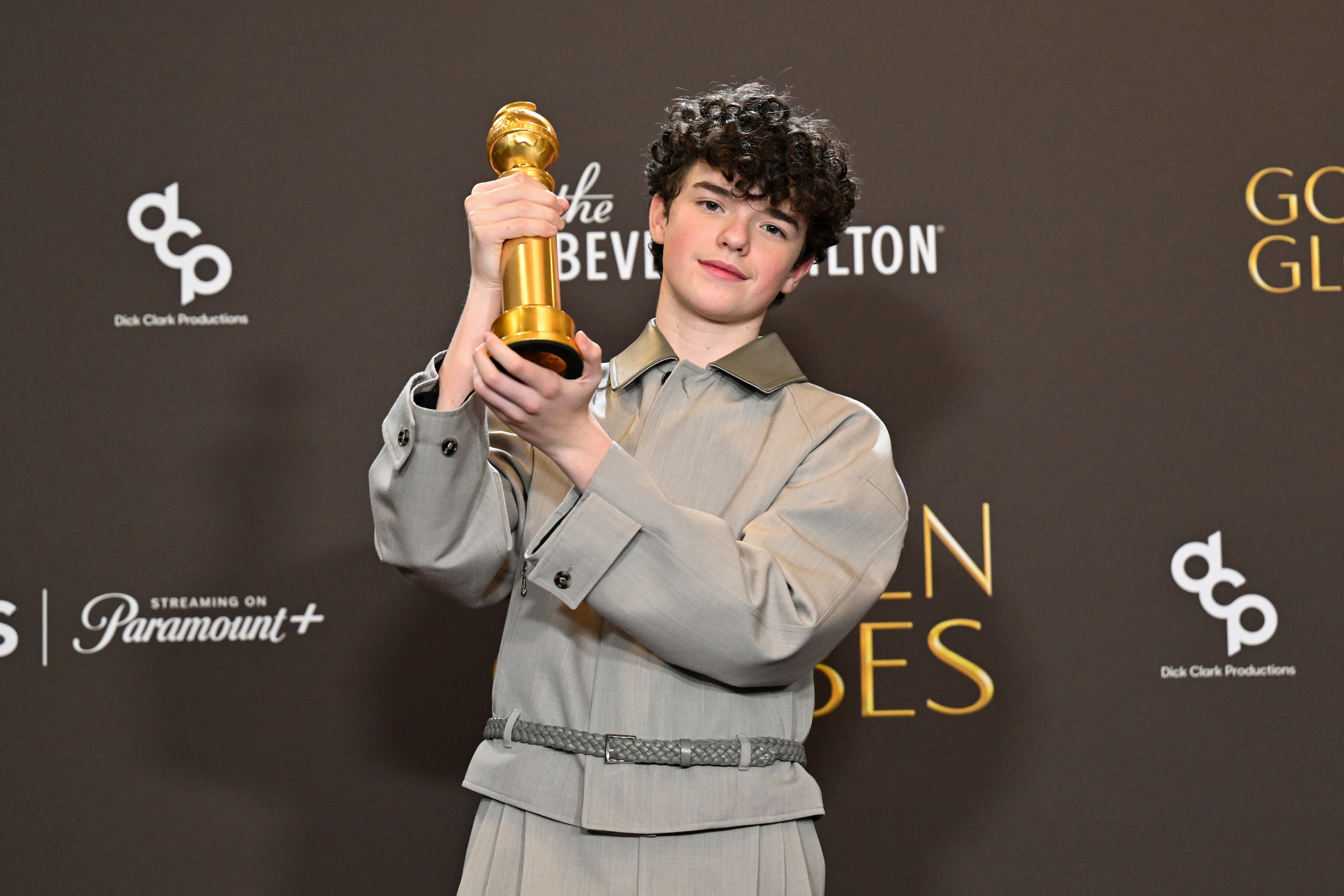 Person successful  grey  outfit holds a Golden Globe award, lasting  successful  beforehand   of a backdrop with lawsuit   logos