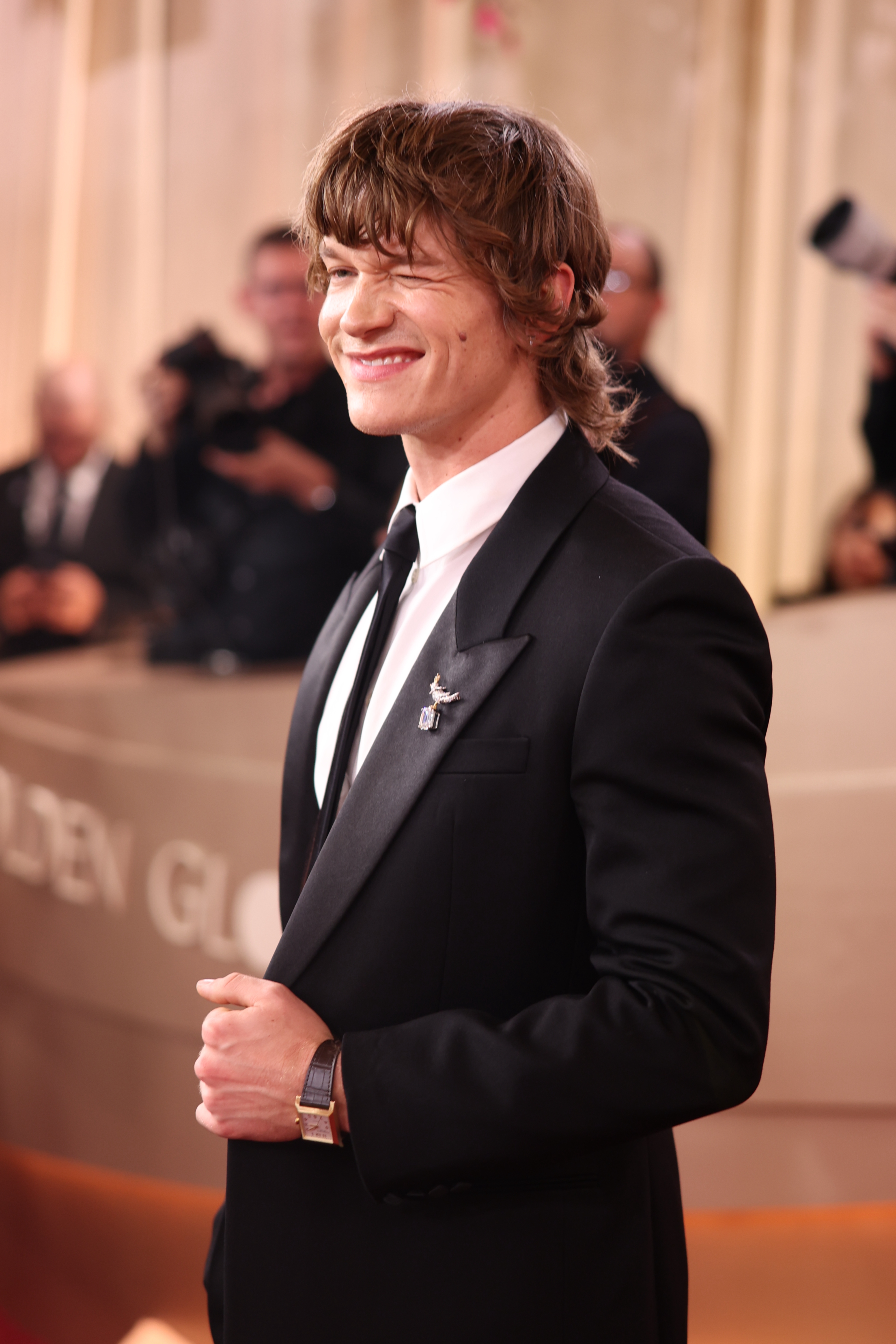 En person i en skreddersydd dress smiler på en formell begivenhets røde løper, der fotografer er synlige i bakgrunnen