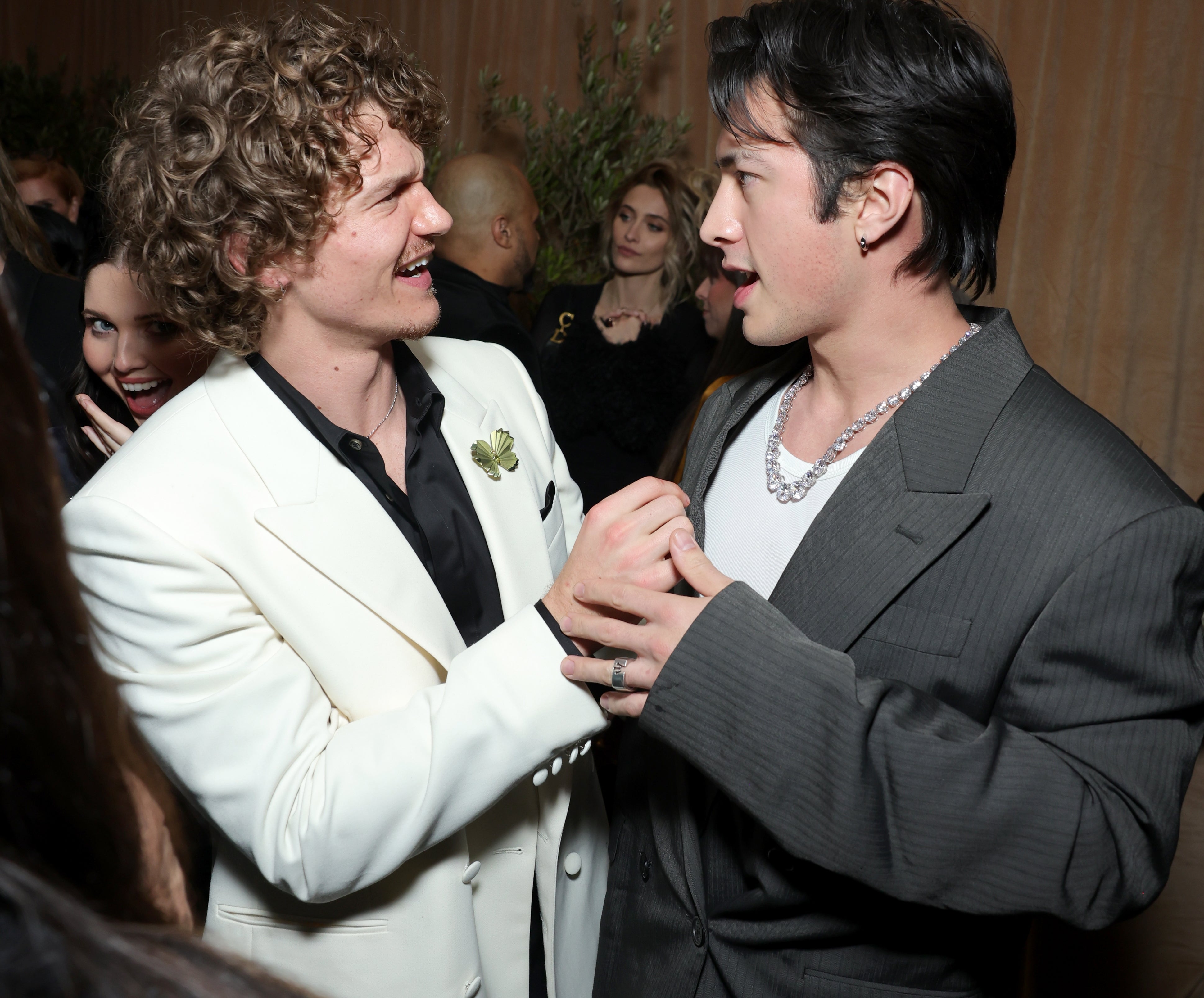 To personer som smiler og holder hender på et arrangement, iført formelle dresser. De ser ut til å nyte en samtale i en sosial setting