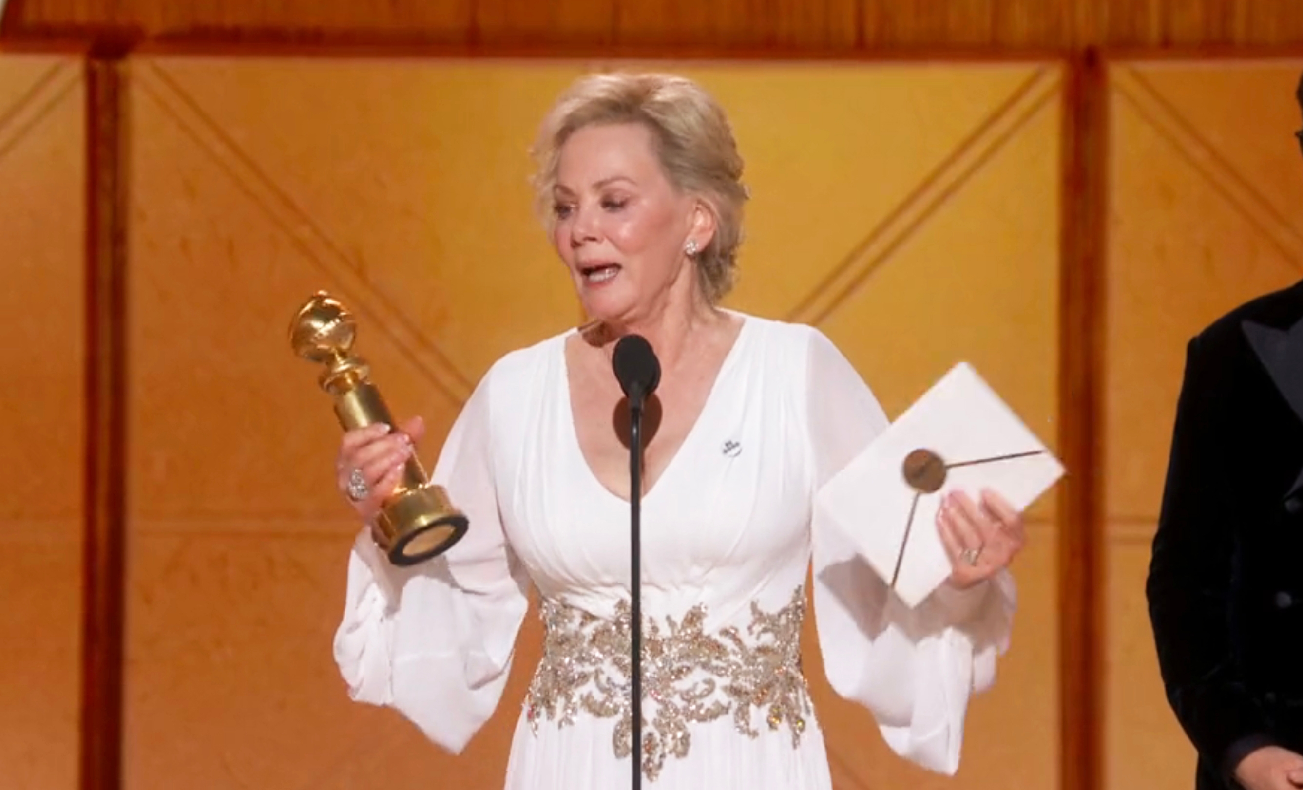 Actor successful  elegant achromatic  gown holding an grant  and envelope connected  stage, speaking into a microphone astatine  an grant  ceremony