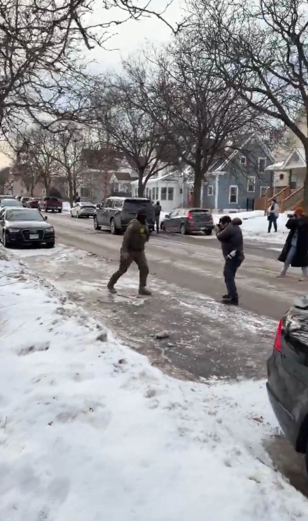 Person går på snødekt gate i vinterantrekk mens en annen person tar bilder. Biler og hus ligger langs gaten, med noen få mennesker i det fjerne