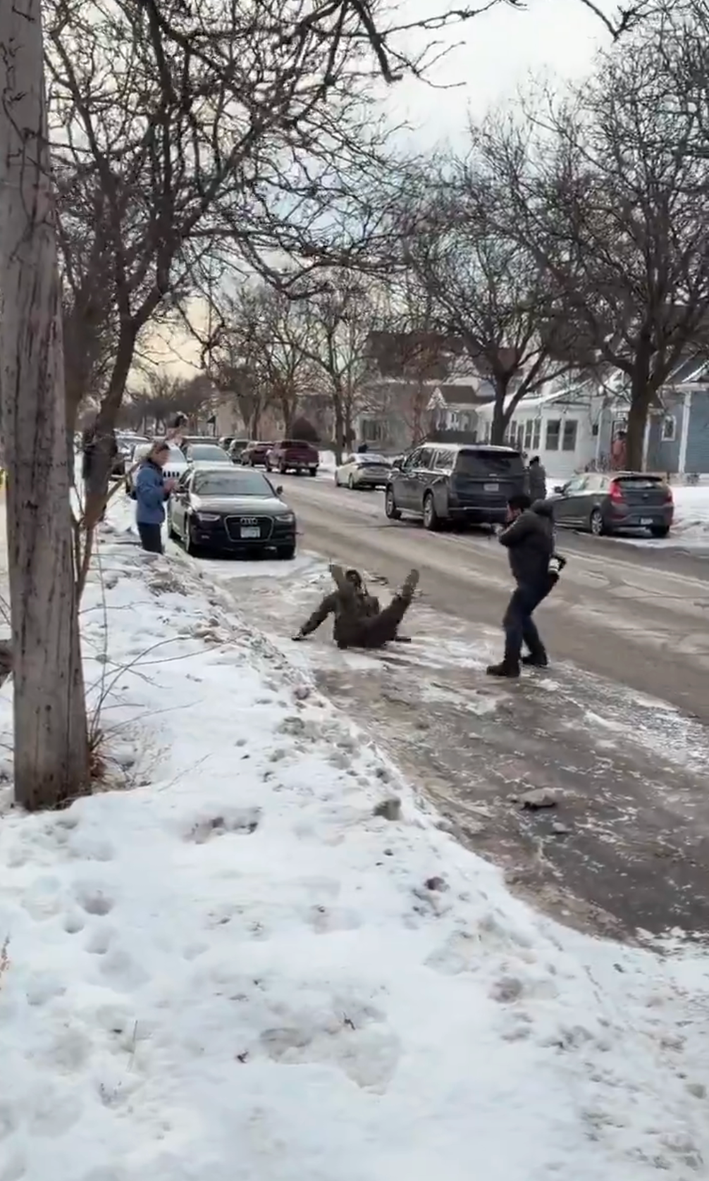 Person som sklir på isete fortau mens to andre i nærheten reagerer. Snødekt gate med parkerte biler og trær i et boligområde