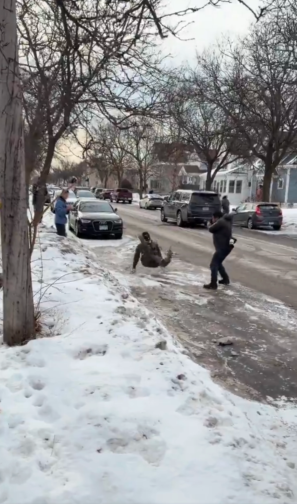 Person glir og faller i en isete nabolagsgate mens andre ser på, noen tar bilder. Snø dekker bakken og parkerte biler står langs gaten