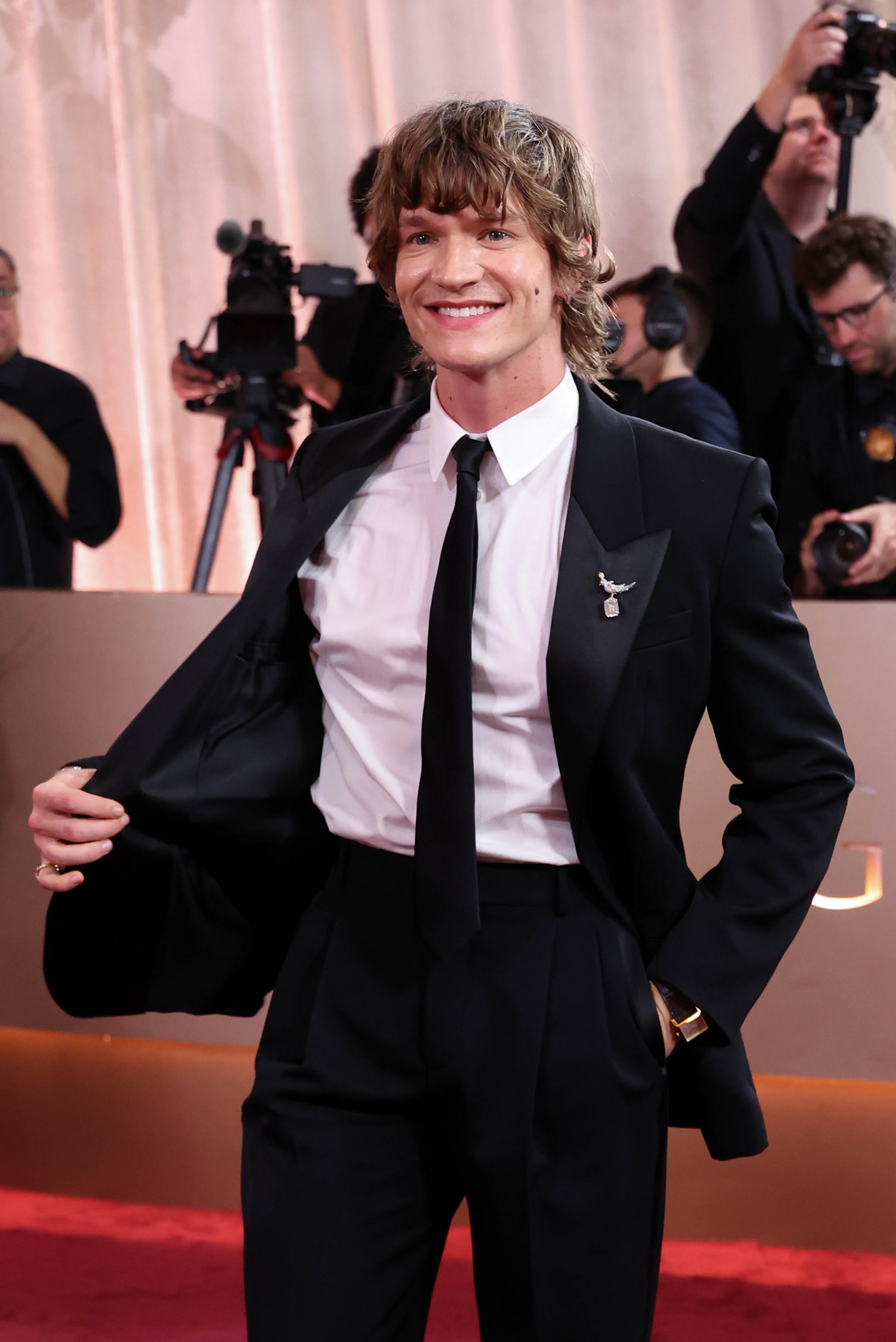 Person i klassisk dress på en rød løper, smiler mens han poserer for bilder. Kameramenn er synlige i bakgrunnen