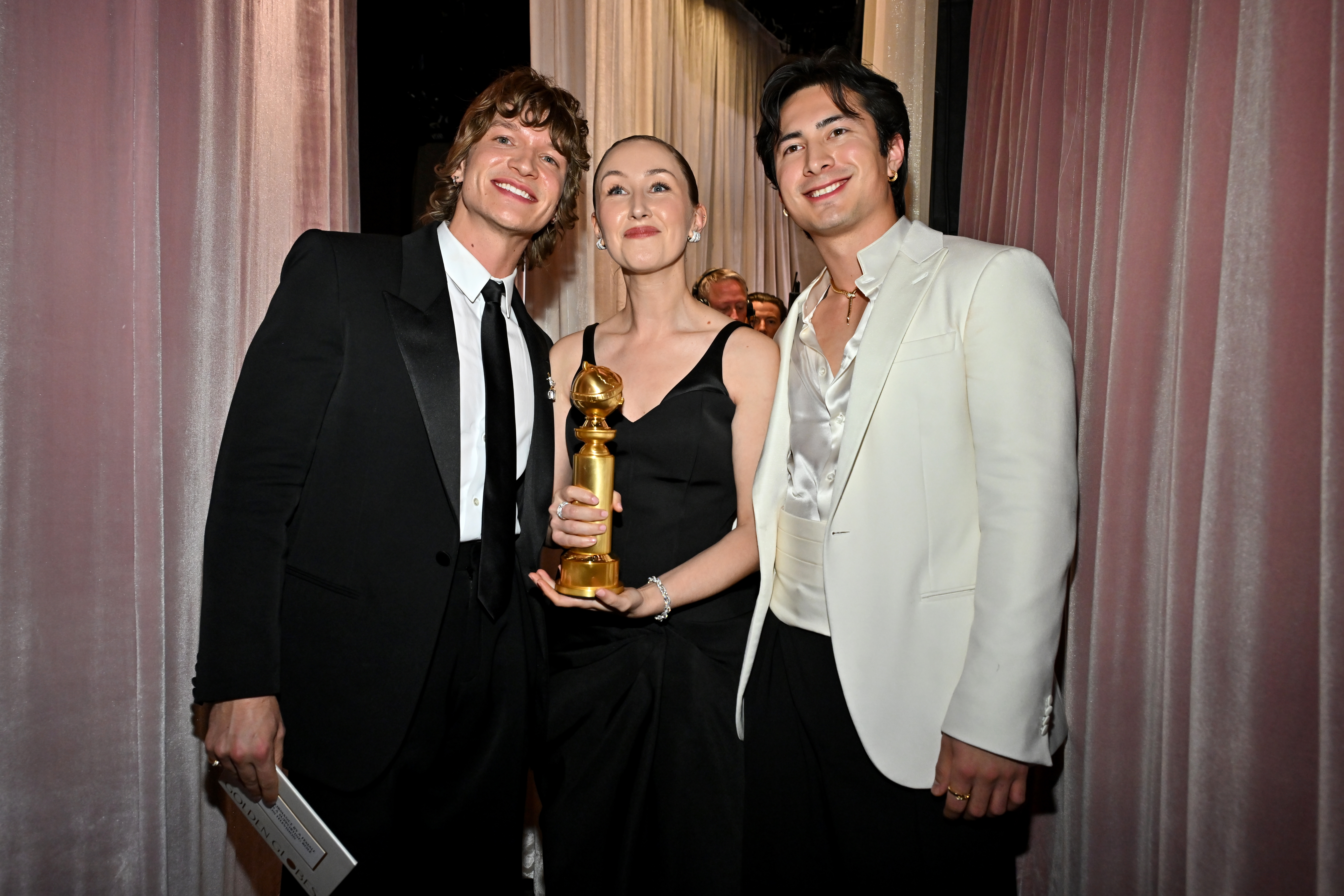 Tre personer poserer sammen på et arrangement. Man holder en gylden pris, smilende. De har på seg formell antrekk: en dress, en kjole og en jakke uten slips