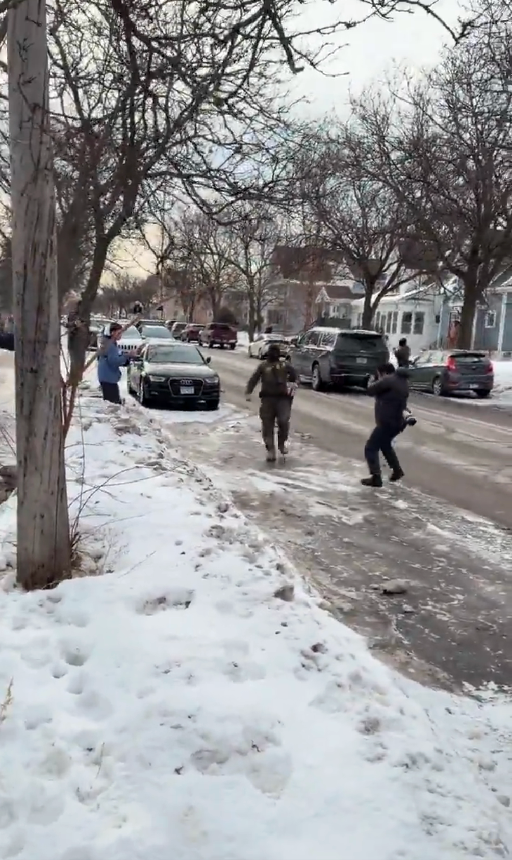 To personer går på en snødekt gate i nærheten av parkerte biler, med nakne trær langs veien en vinterdag