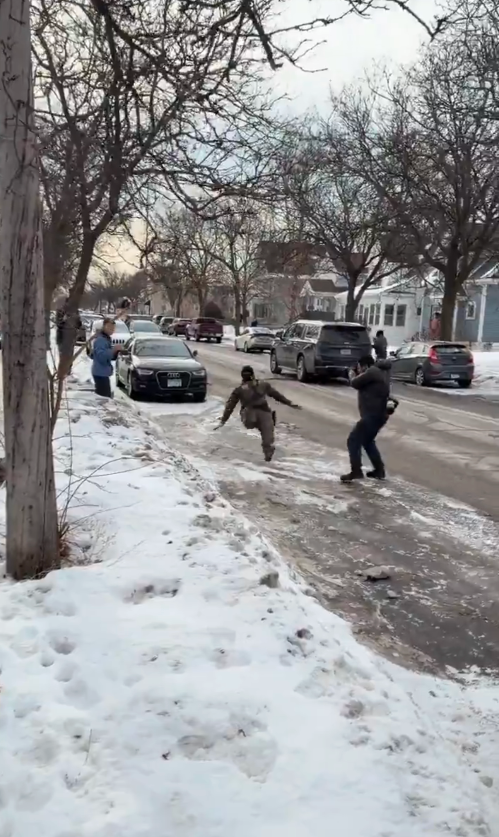 Person i vinterutstyr glir nedover en isete gate mens tilskuere ser på og registrerer, og fremhever de farlige værforholdene