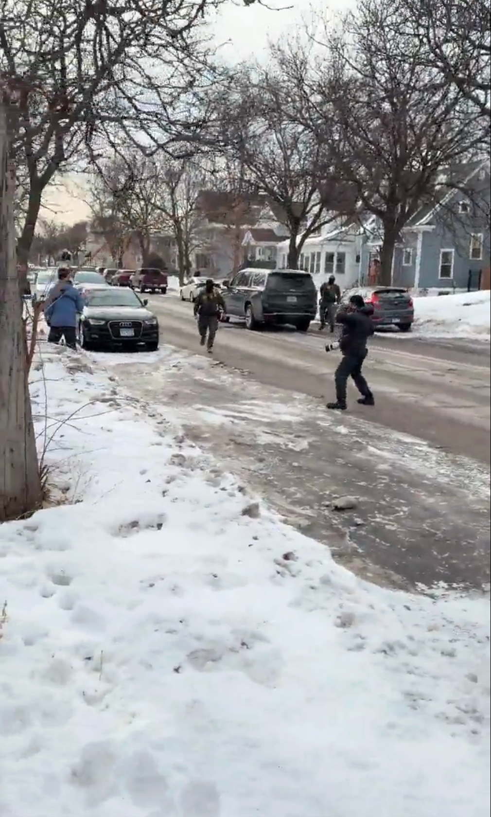 Folk går forsiktig i en snødekt gate med parkerte biler, sannsynligvis etter et nylig snøfall. Hus og trær langs gaten
