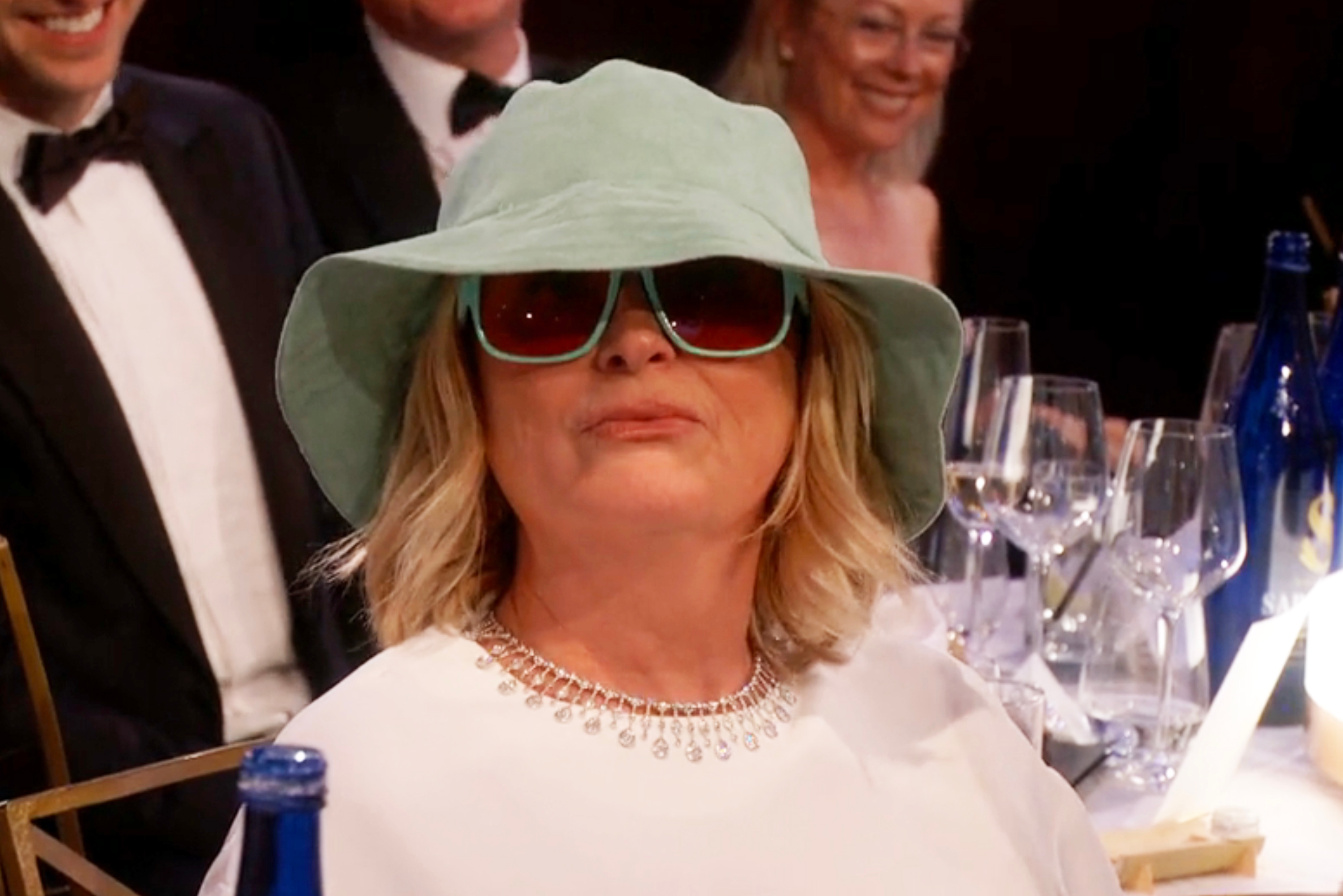 Person sitting astatine  a ceremonial  lawsuit   wearing a wide-brimmed chapeau  and sunglasses, dressed successful  a achromatic  outfit with a decorative neckline
