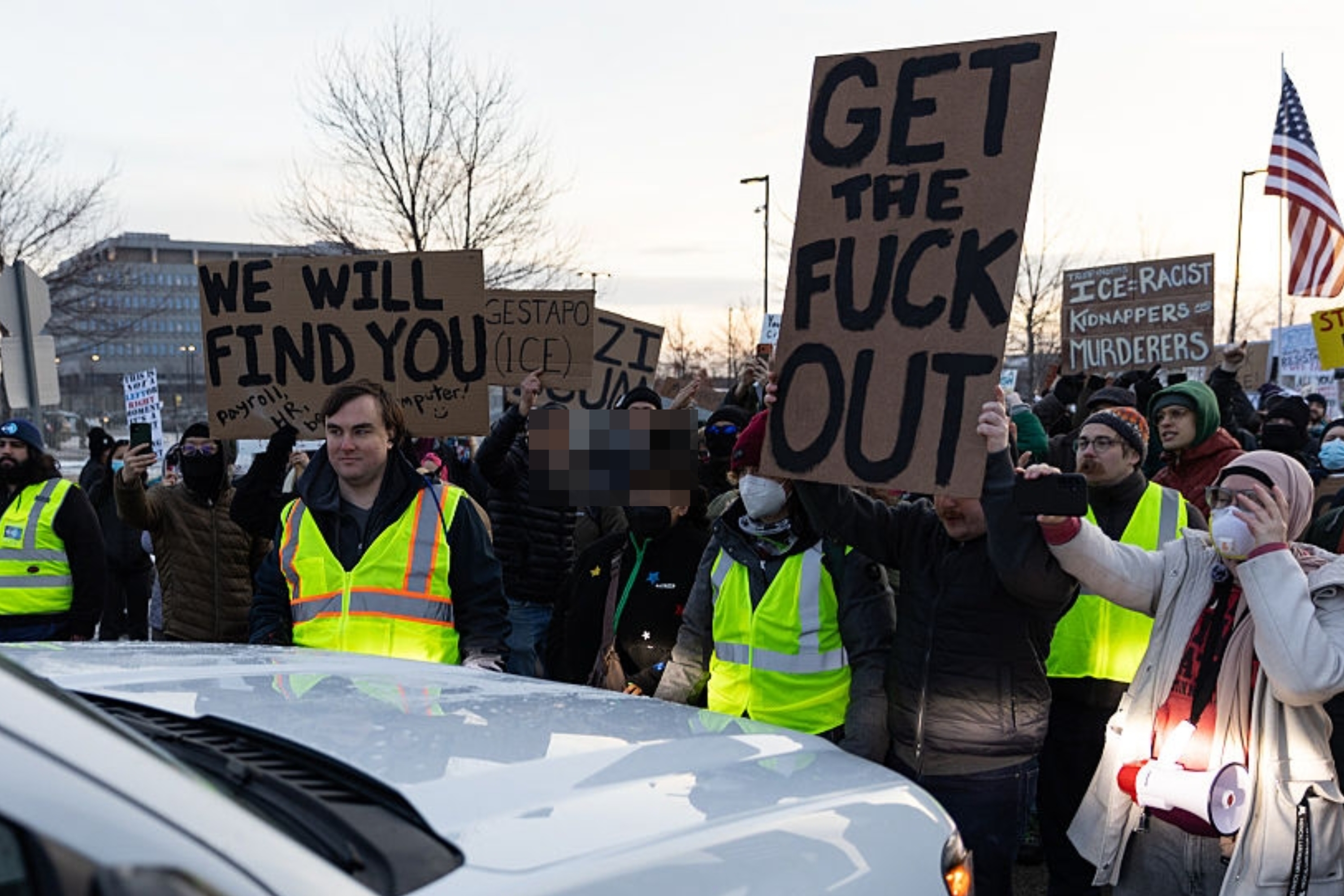 Demonstranter samles som holder skilt; man leser "Få ut (ekspletive) ut." Noen bruker refleksvester. Det er et kjøretøy i forgrunnen