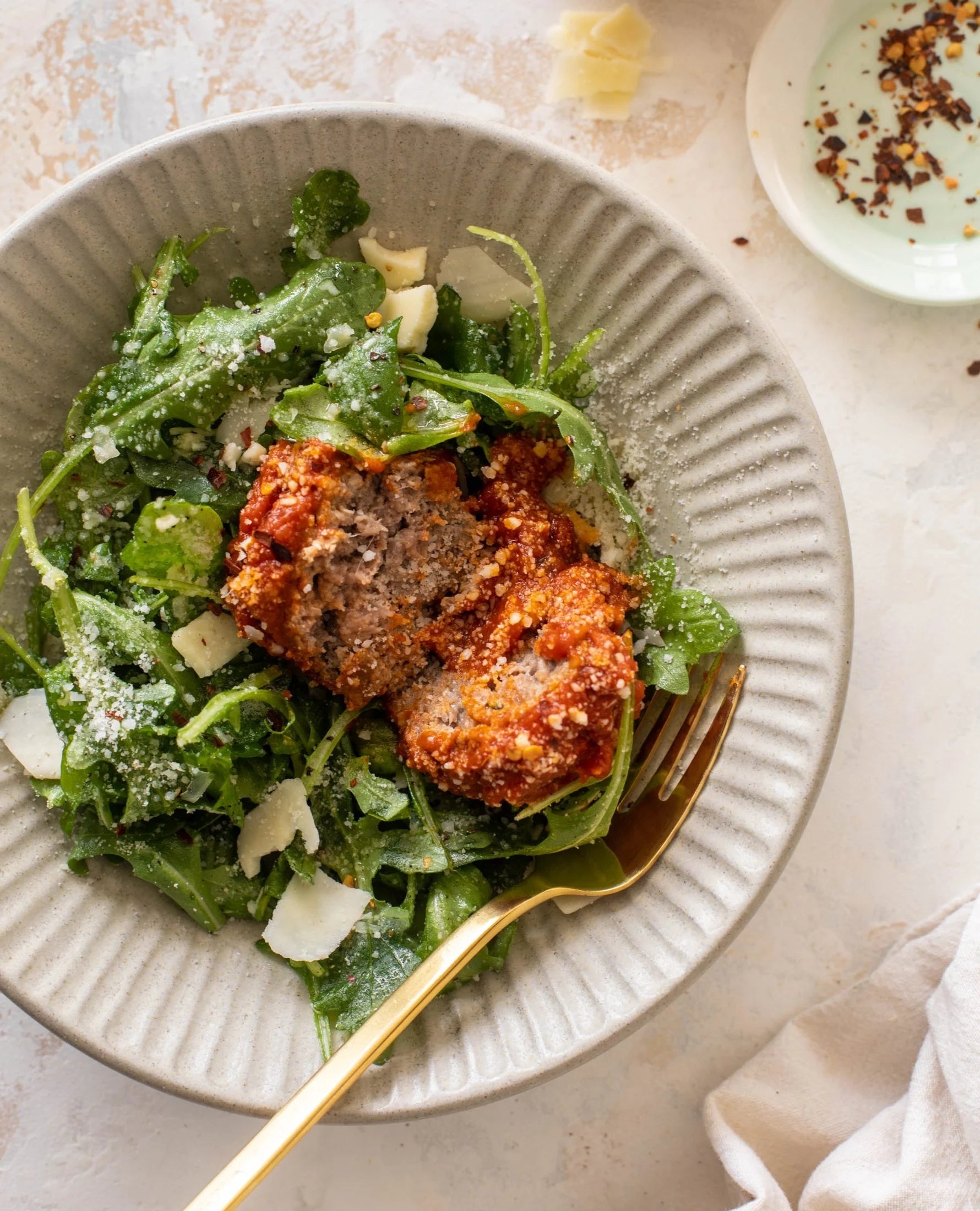 A plate with leafy greens and a saucy meatball topped with grated cheese, paired with a gold fork on a textured dish