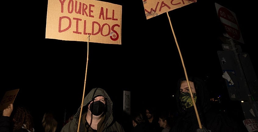 To demonstranter holder skilt med teksten «DU ER ALLE DILDOS»; og "ICE IS WACK" på en nattlig demonstrasjon