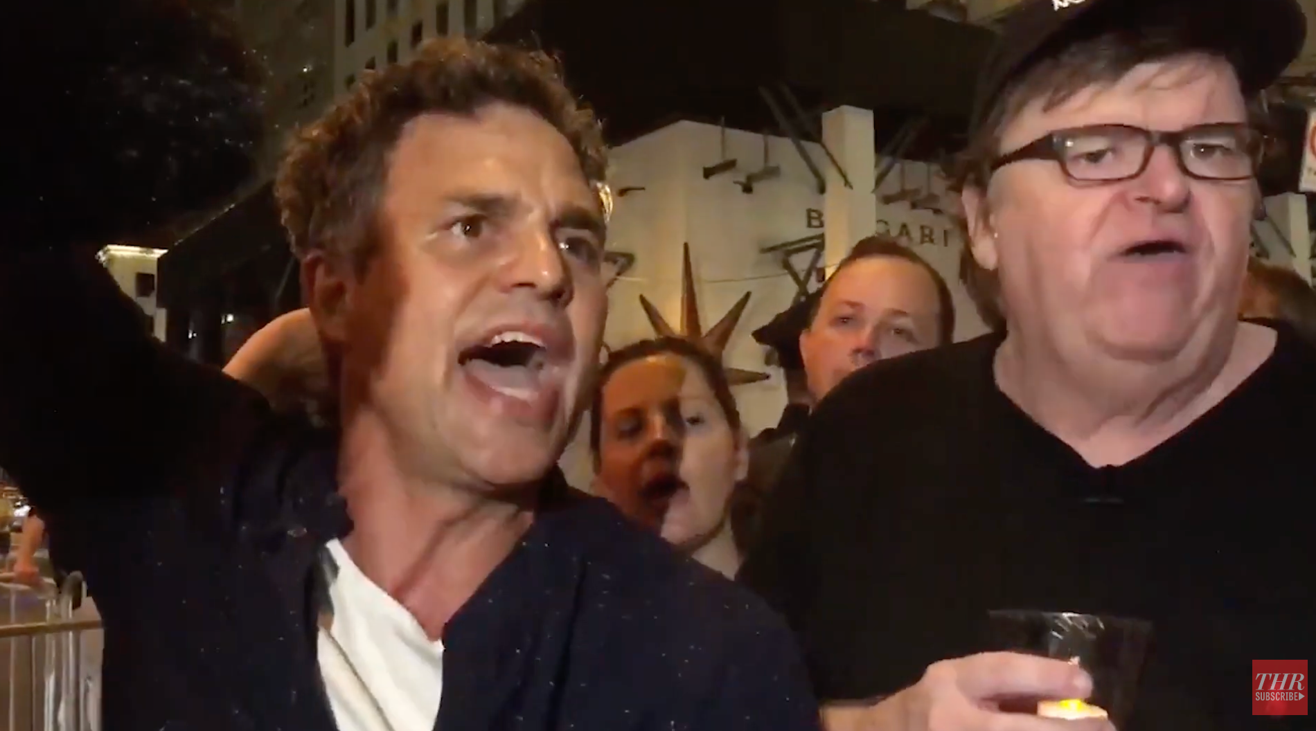 To personer snakker lidenskapelig foran en folkemengde på et utendørsarrangement. Den ene har på seg en uformell skjorte, den andre en caps og briller og holder et stearinlys