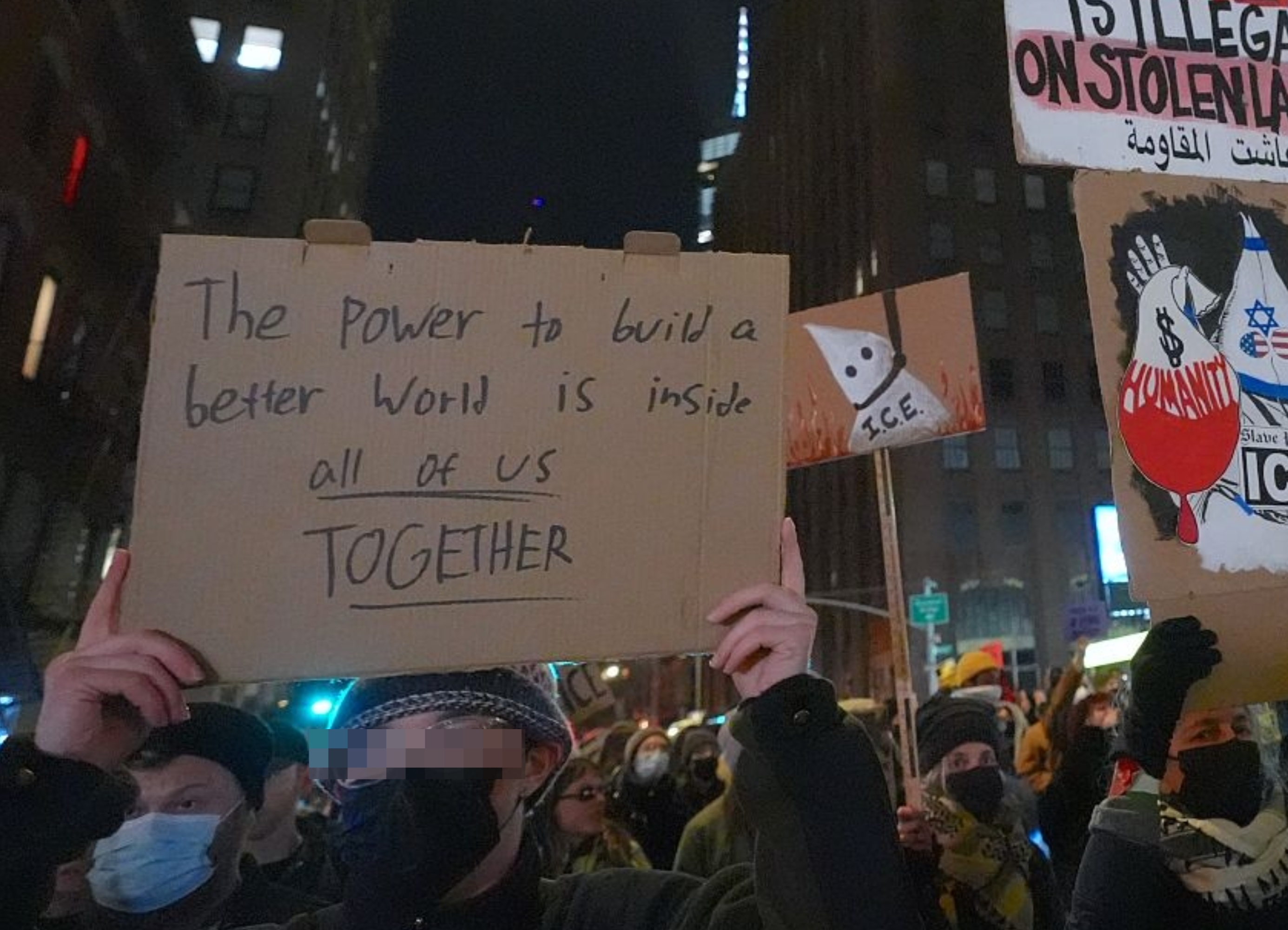 Folk på en protest holder skilt. Signer meldinger: "Kraften til å bygge en bedre verden er inne i oss alle SAMMEN" og "Ingen er ulovlig på stjålet land."