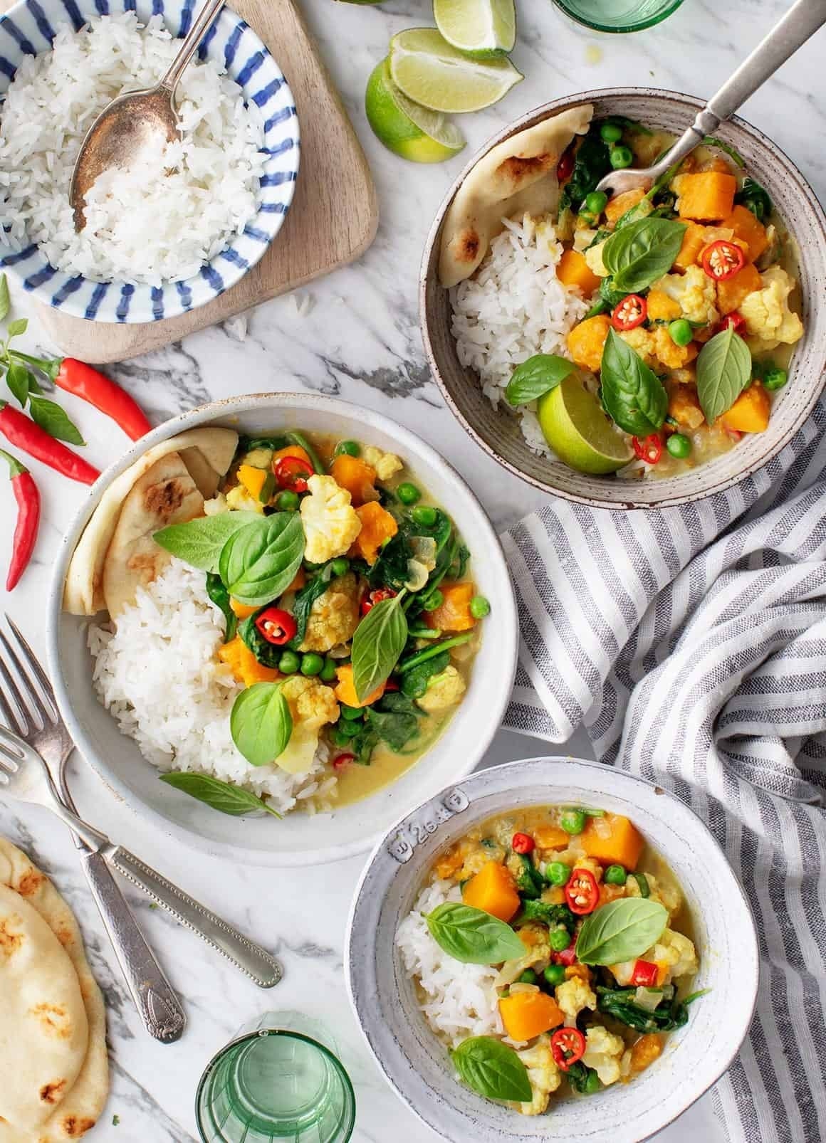 Plates of vegetable curry with rice and naan, garnished with basil and chili slices, set on a marble surface with lime wedges and fresh chili peppers