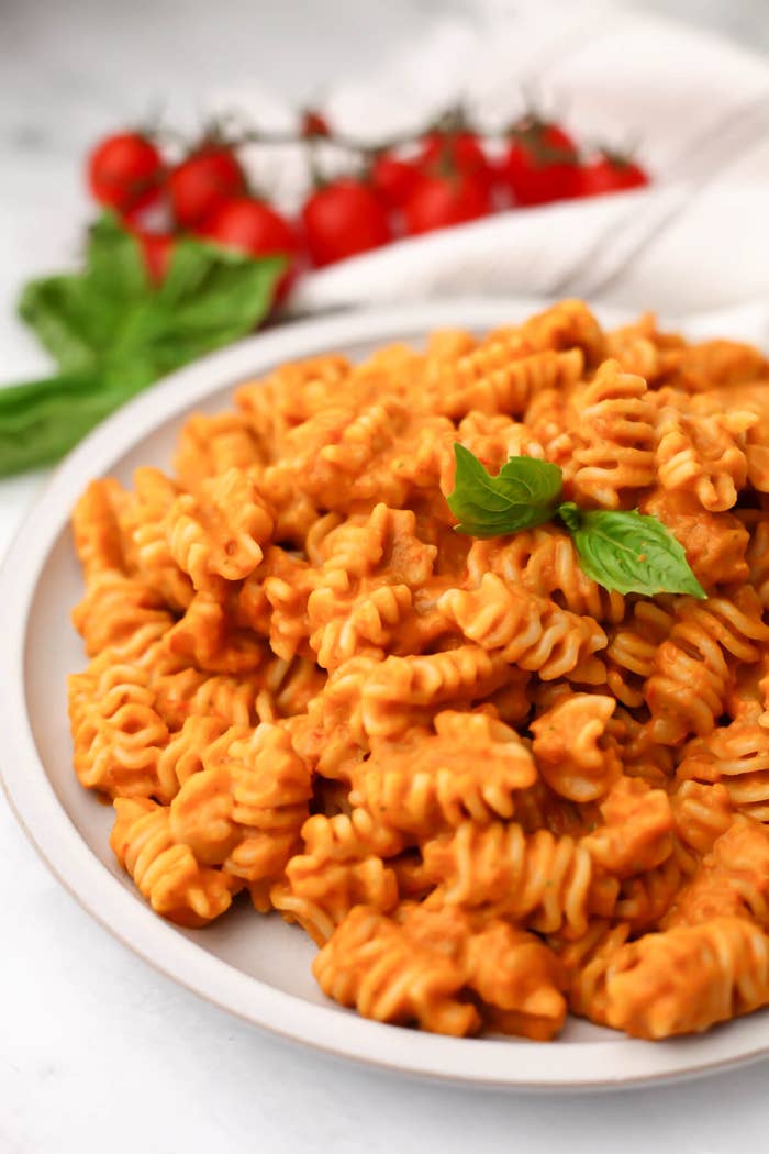 A plate of cooked rotini pasta topped with a creamy sauce and a basil garnish; tomatoes and basil in the background