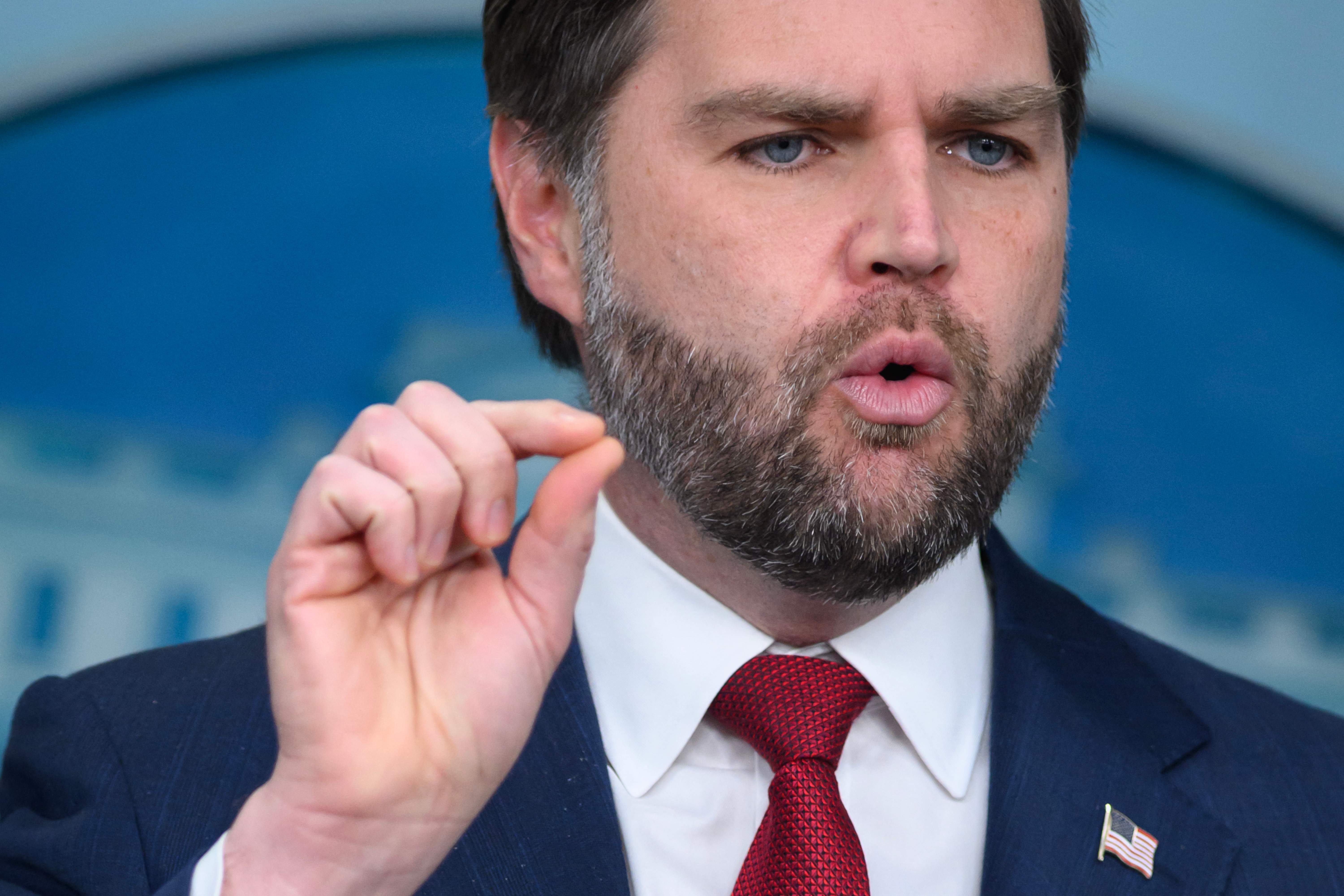 En mann i dress og rødt slips snakker ettertrykkelig, holder hånden opp i en gest, med et uskarpt regjeringslignende bakteppe bak seg