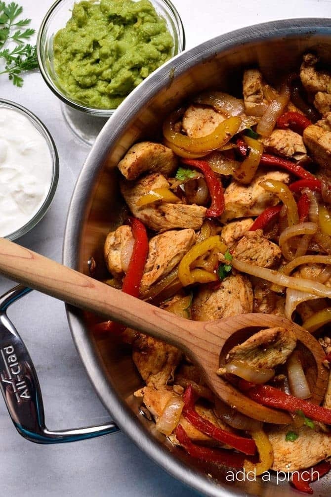 Chicken fajitas with bell peppers and onions in a pan, with sides of guacamole and sour cream beside it