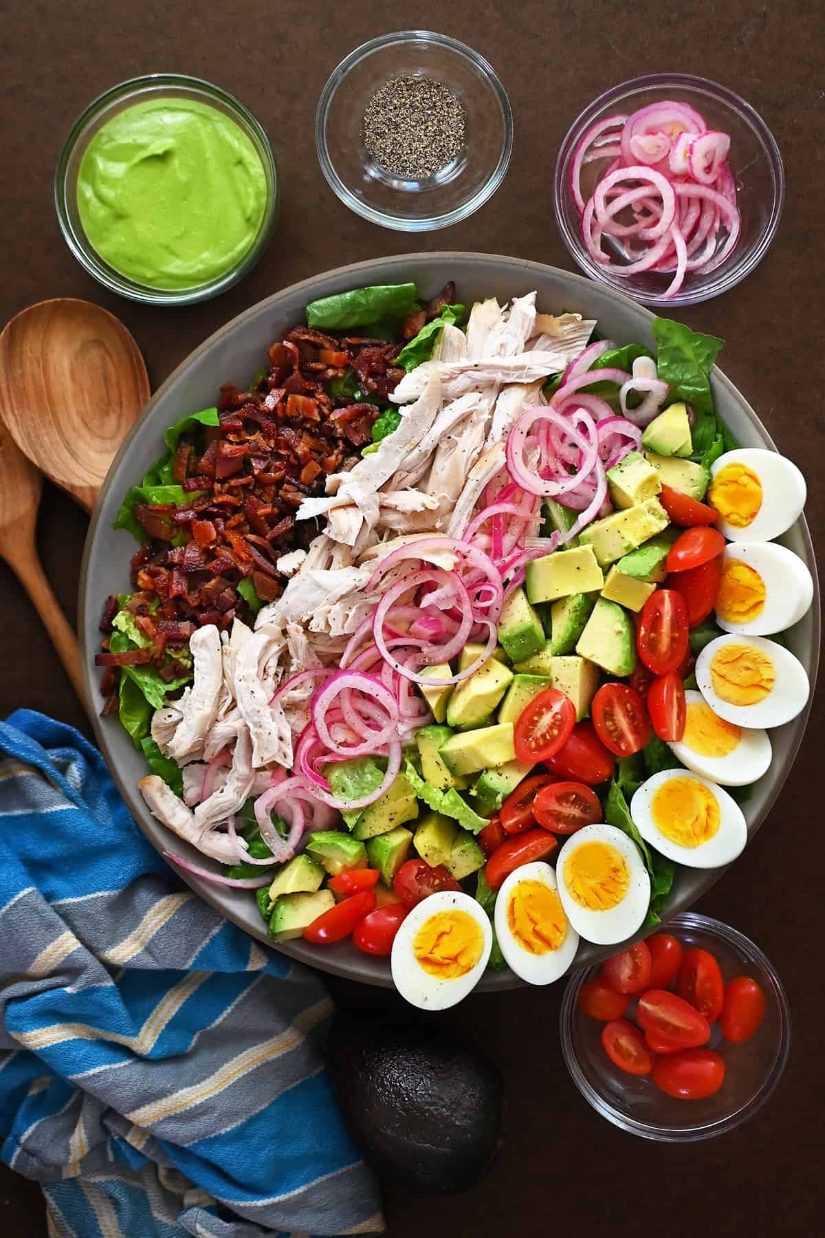 A large salad is presented with ingredients in sections: sliced chicken, boiled eggs, avocado, cherry tomatoes, bacon, and red onions, surrounded by dressing bowls
