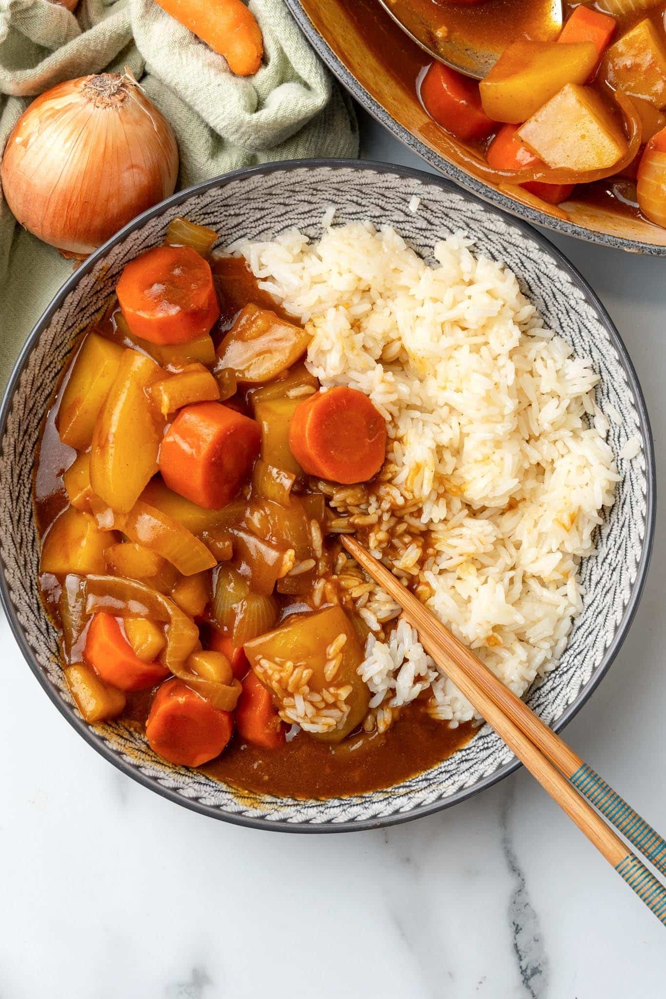 A bowl of Japanese curry with carrots and potatoes served with rice, accompanied by chopsticks, placed on a marble surface