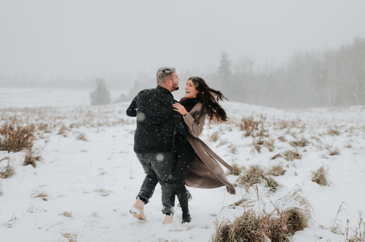 A couple joyfully dances in a snowy field, surrounded by gentle snowfall and winter grasses