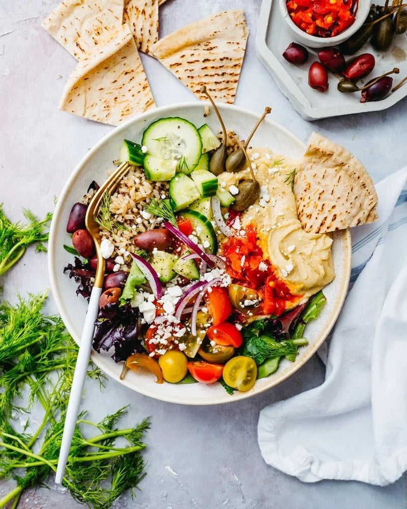 A Mediterranean salad with cucumbers, tomatoes, olives, red onions, feta, and hummus, garnished with pita slices and fresh dill