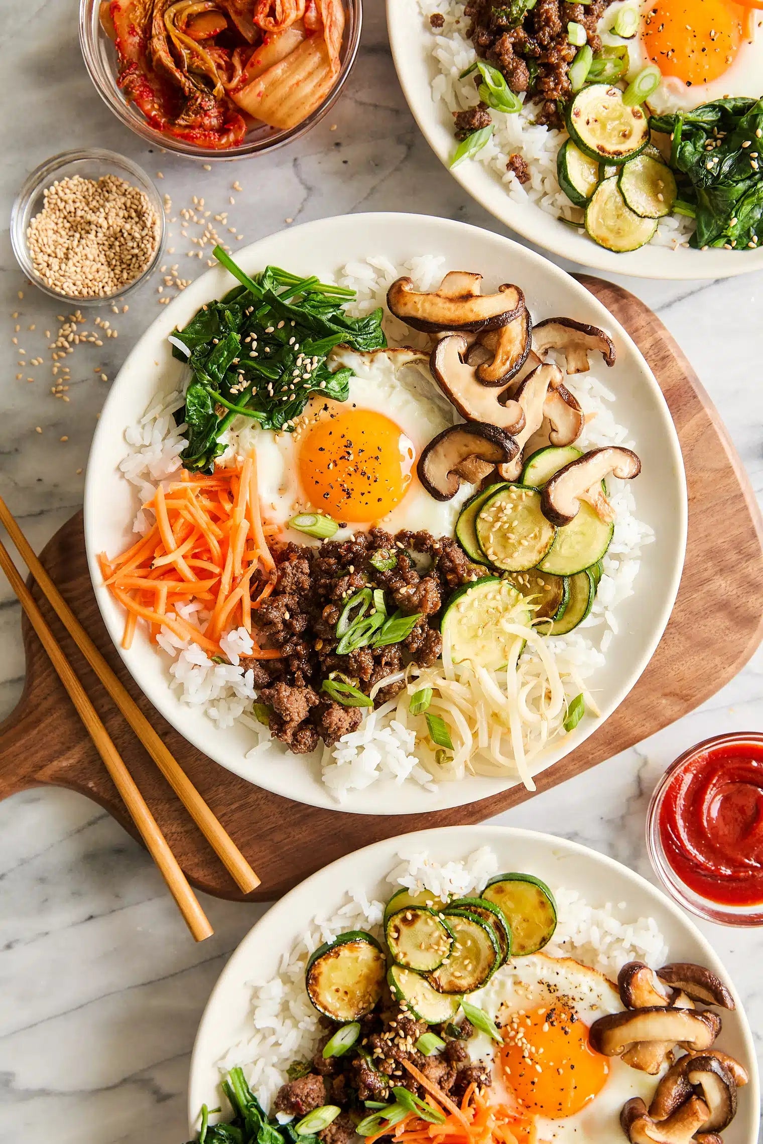 Three bowls of bibimbap with beef, rice, vegetables, and fried eggs, accompanied by kimchi, sesame seeds, and sauce on the side. Chopsticks nearby