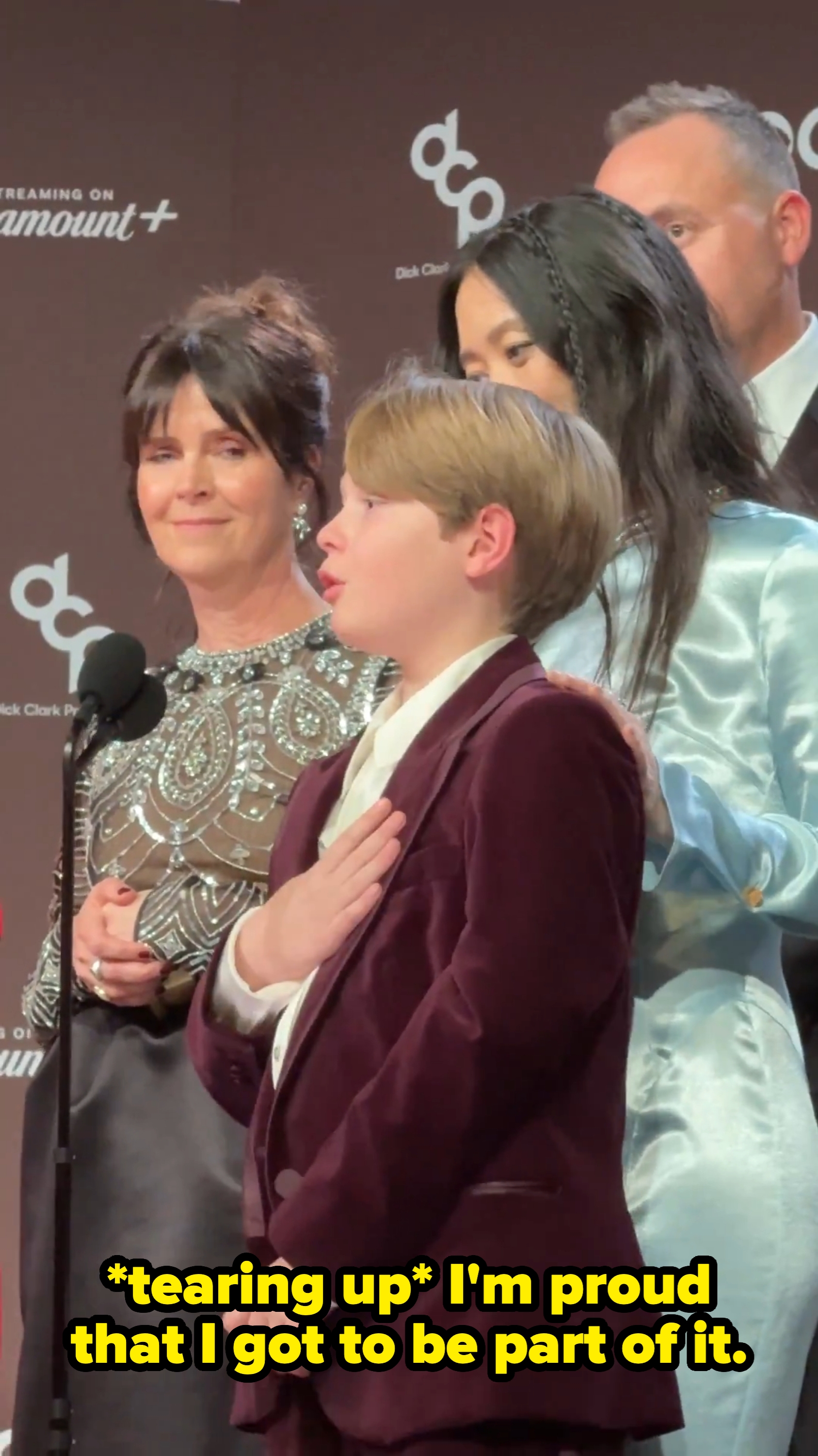 A kid wearing a velvet suit speaks astatine a microphone. Three adults basal behind, dressed successful ceremonial attire, smiling successful support