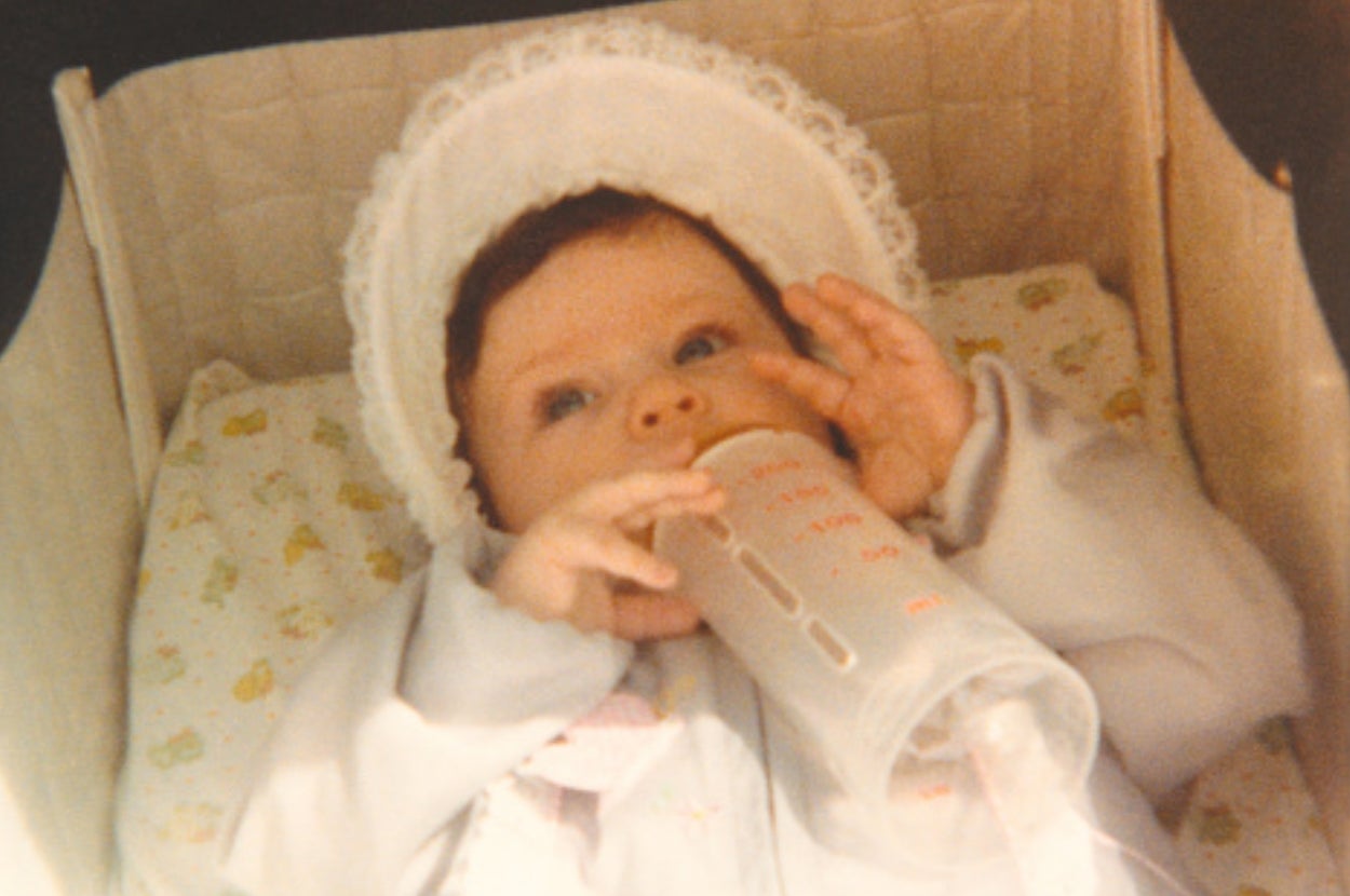 Baby in a bonnet holding a bottle while lying in a cradle