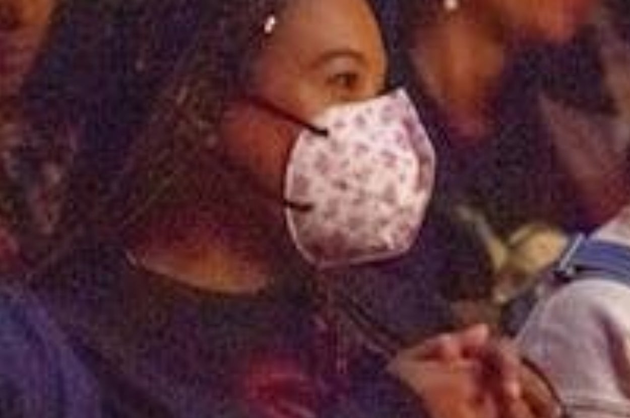 Person wearing a patterned mask, sitting in a crowd, appearing attentive. Others around are also focused in the same direction