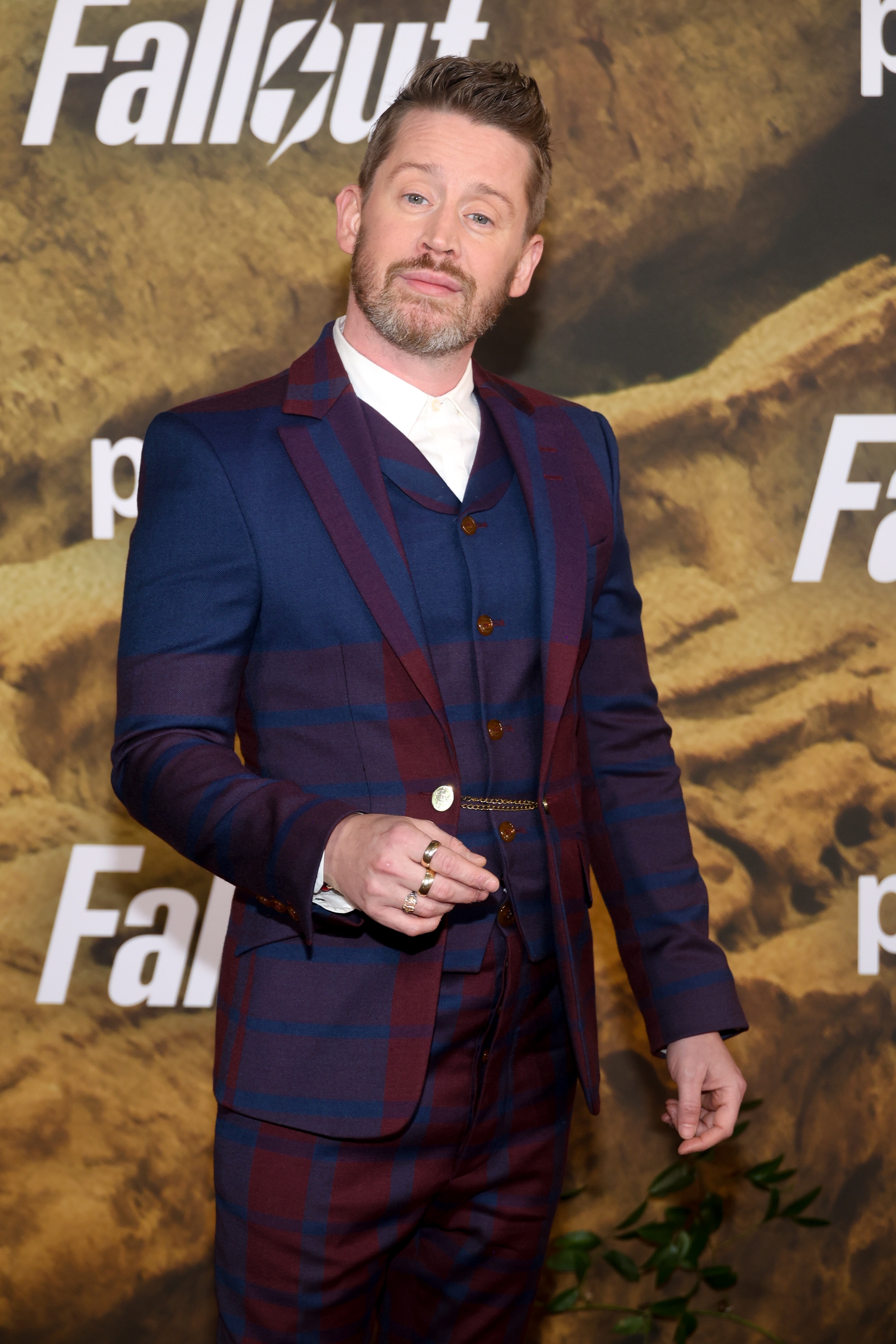 Person in a tailored plaid suit poses confidently on a media event backdrop with "Fallout" text
