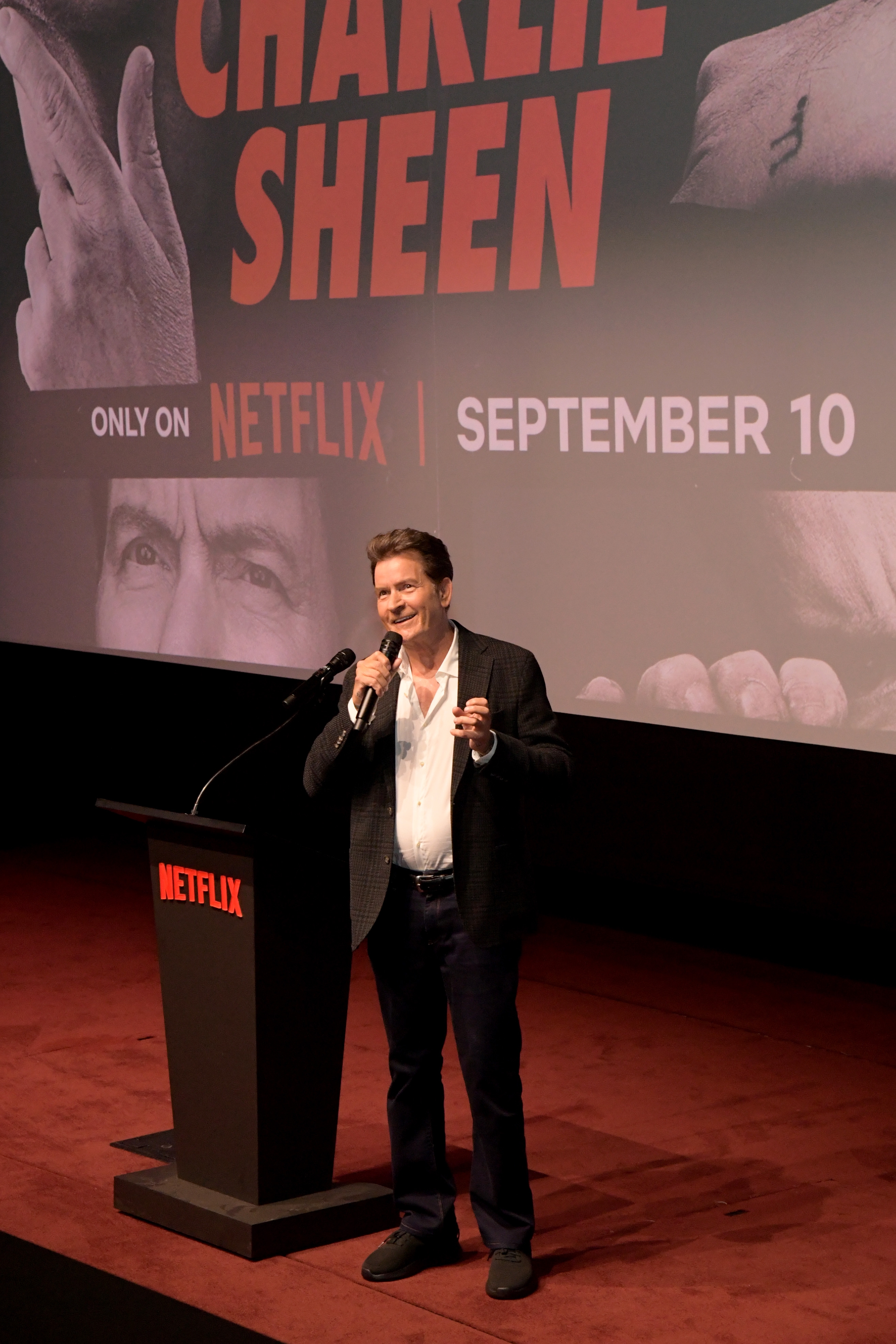 Celebrity on stage at a Netflix event, wearing a suit jacket and white shirt with an image of a film poster in the background