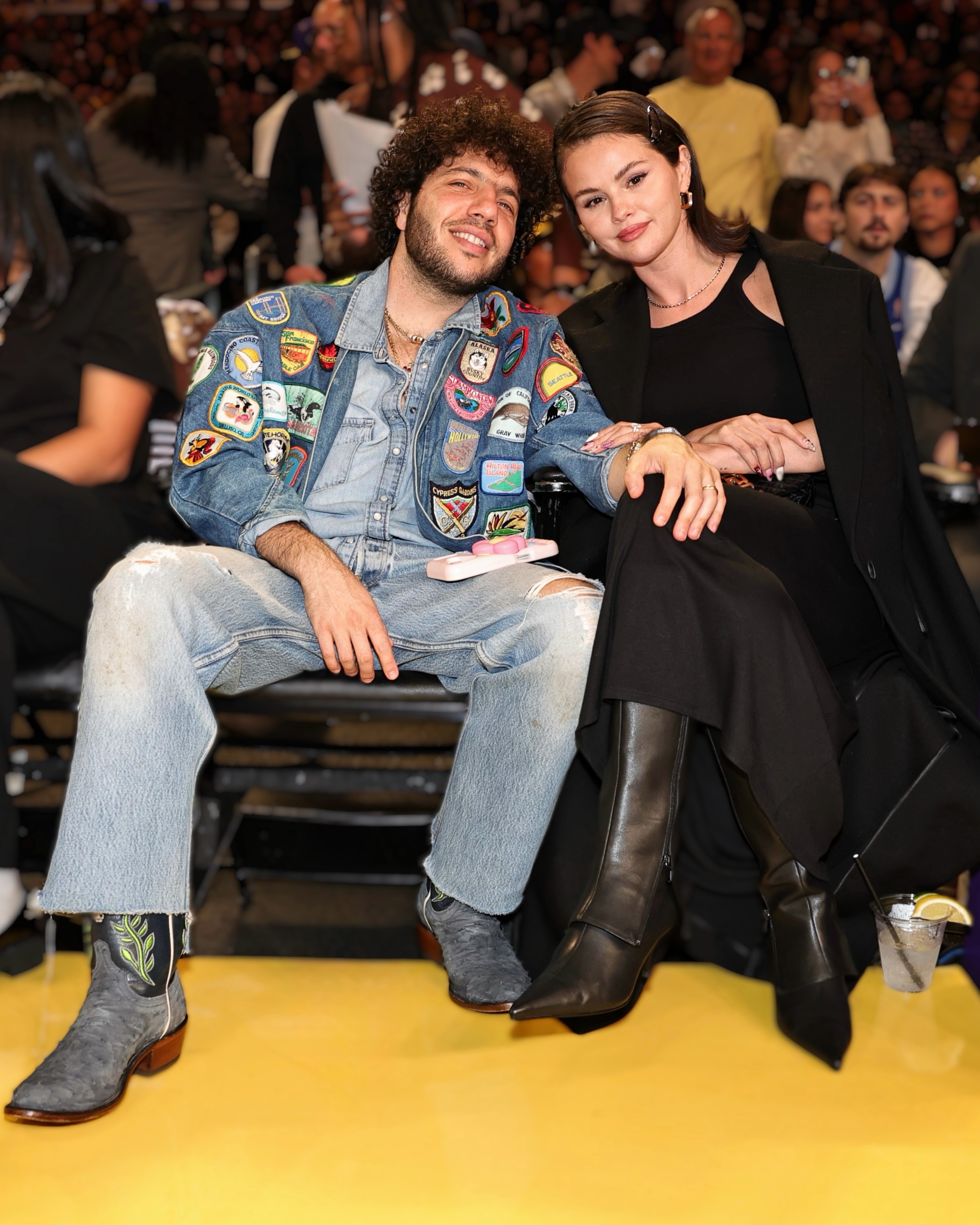 Two people sit courtside. One wears a patch-covered denim jacket and jeans, and the other wears a black dress and knee-high boots. They pose casually