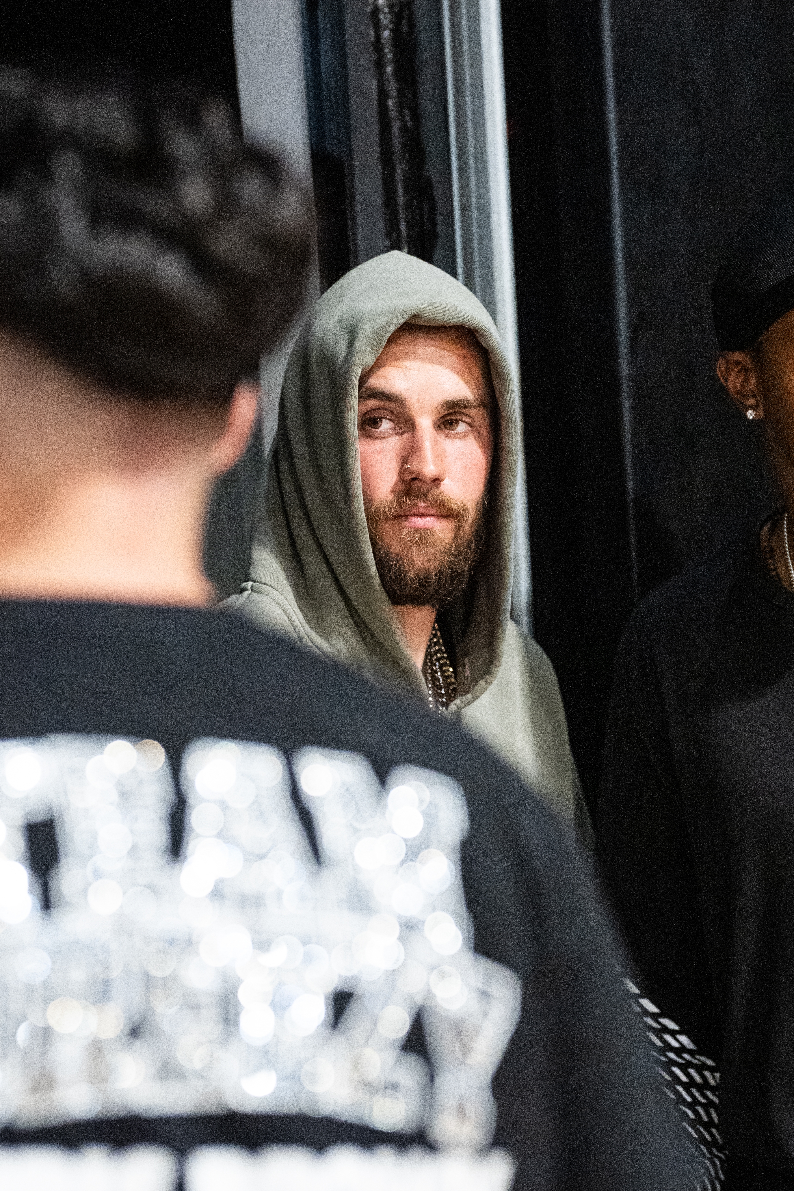 A bearded man in a hoodie stands indoors, looking towards another person. They appear to be in conversation at a casual event