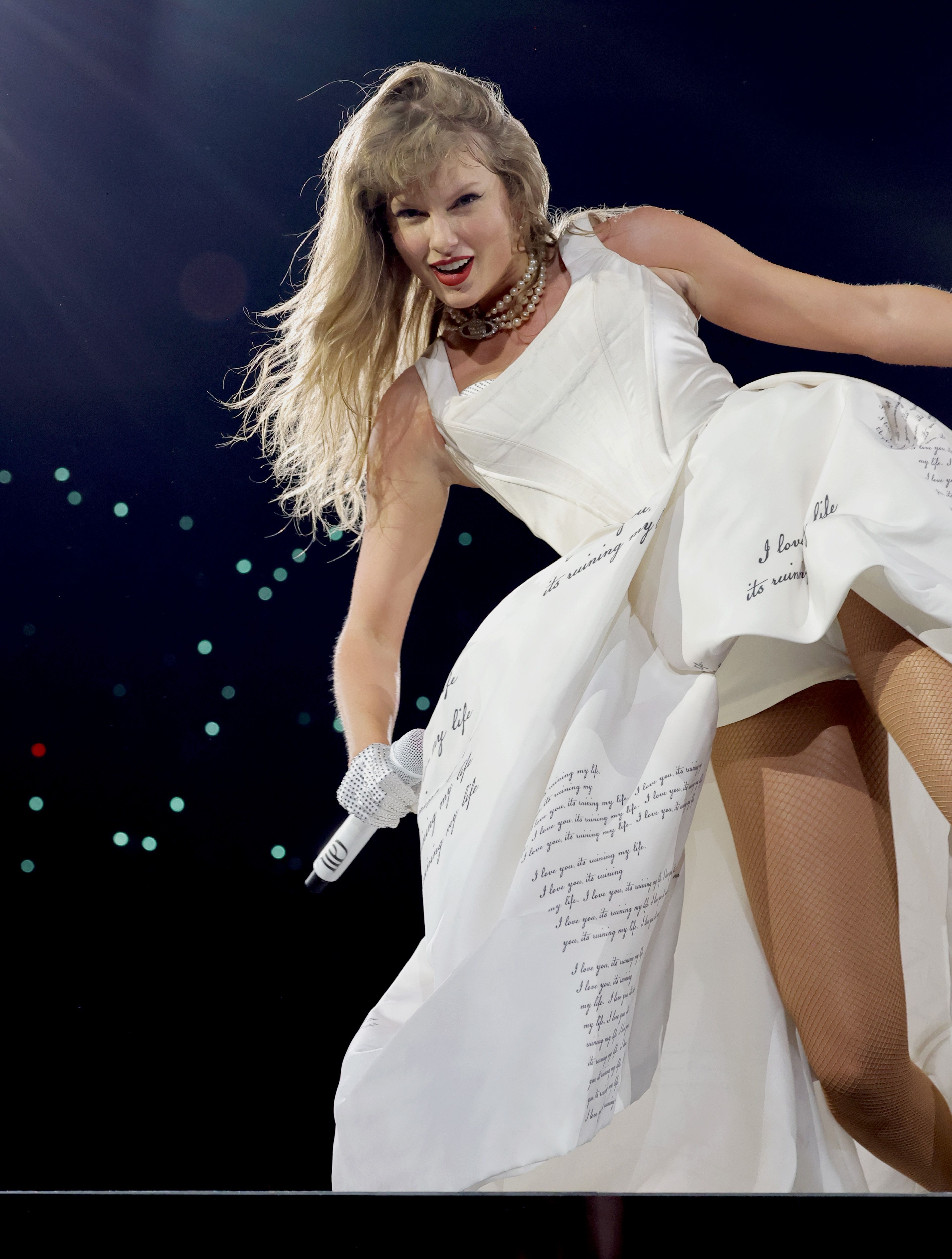 Singer in a white dress with handwritten text, performing energetically on stage with a microphone, surrounded by lights