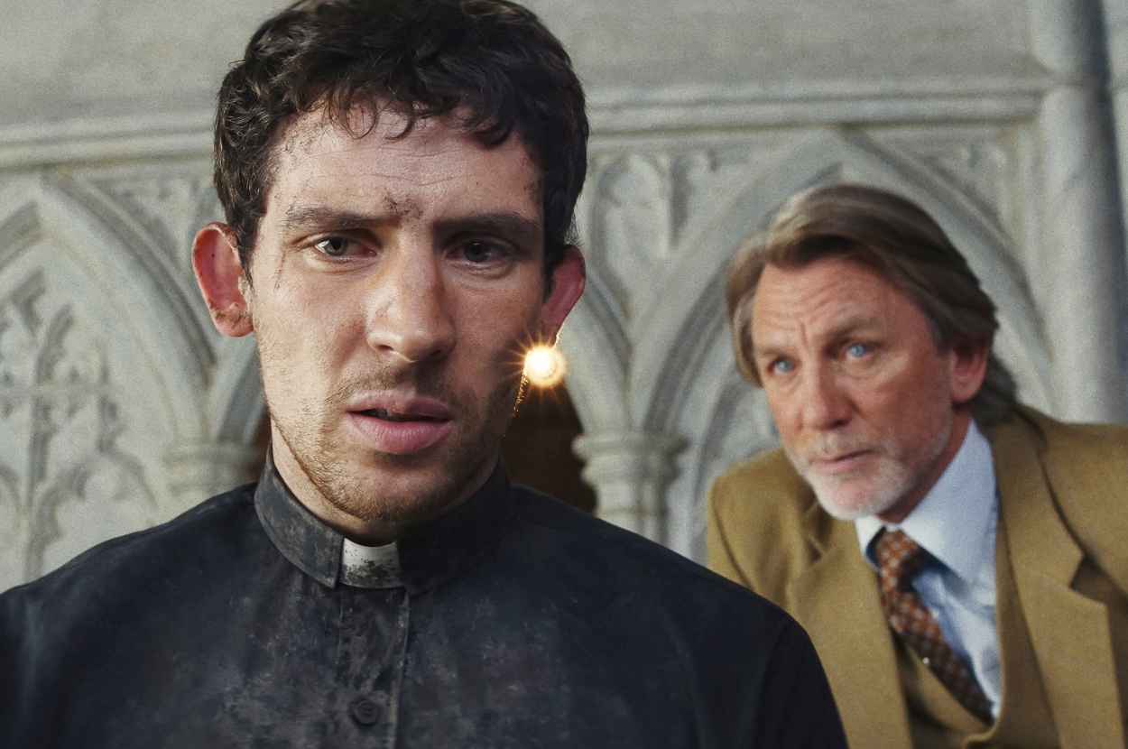 A worried man in a priest's collar is in focus, with a concerned older man in a suit behind him in a gothic setting