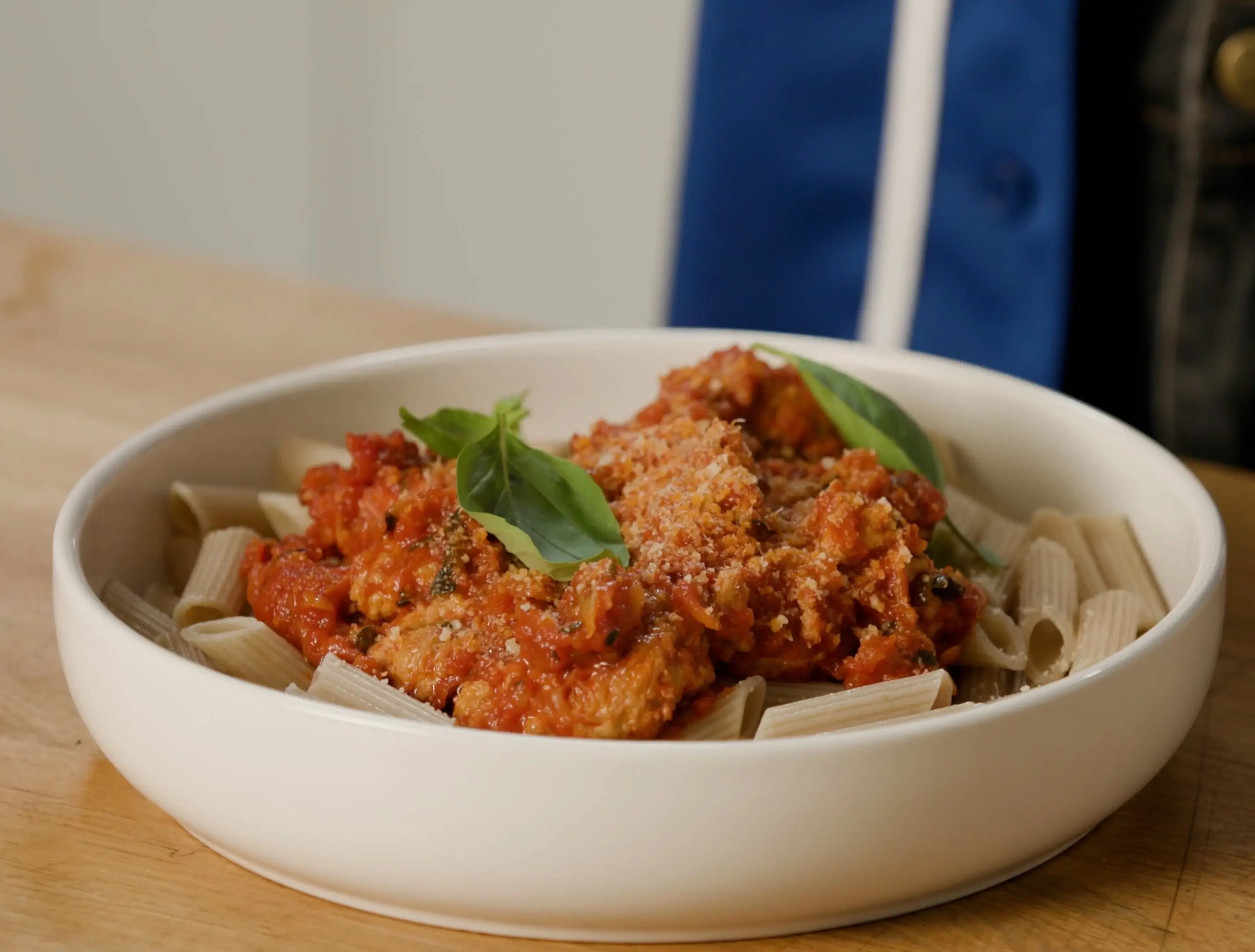 A bowl of pasta topped with tomato sauce, sprinkled with cheese, and garnished with basil leaves, placed on a wooden table