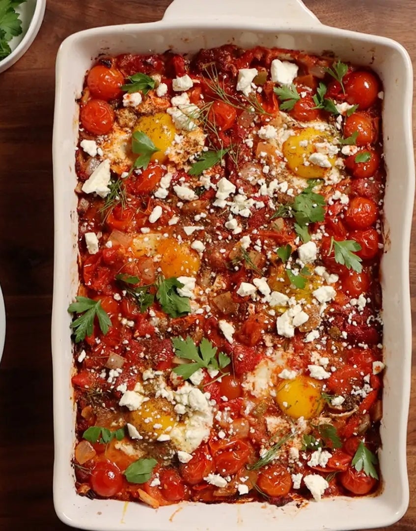 Baked shakshuka in a dish with eggs, tomatoes, and feta, garnished with parsley. Served with pita bread and herbs on the side