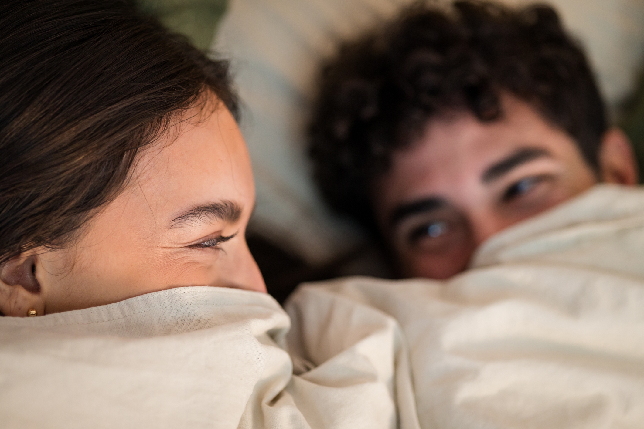 A couple playfully lies under bed covers, smiling at each other, conveying intimacy and affection