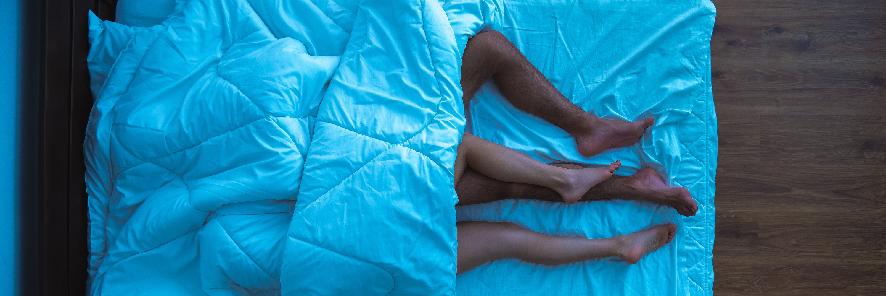Overhead view of a couple's legs intertwined under a blanket in bed