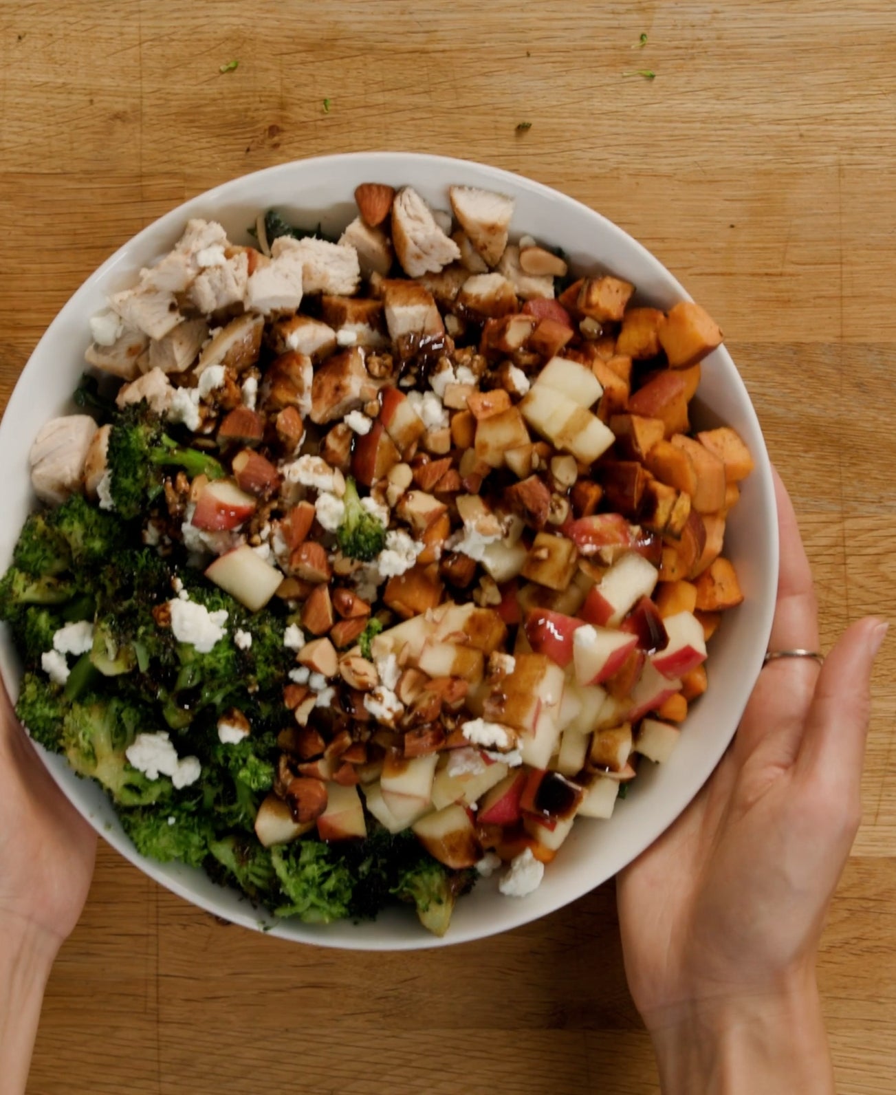 Person holding a bowl of salad with chicken, apples, sweet potatoes, greens, cheese, and nuts on a wooden surface
