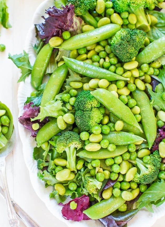 A fresh salad on a white plate, featuring snap peas, broccoli florets, green peas, edamame, and mixed leafy greens