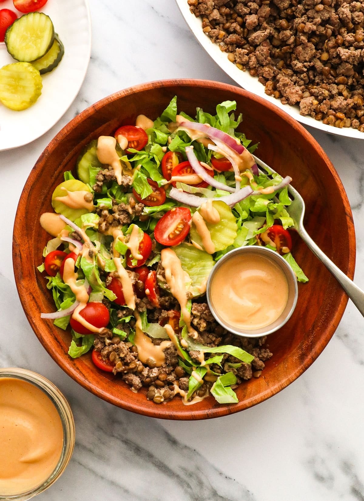 Salad with lettuce, cherry tomatoes, onion, and ground beef, drizzled with sauce, in a wooden bowl. Small bowls of dressing and pickles nearby