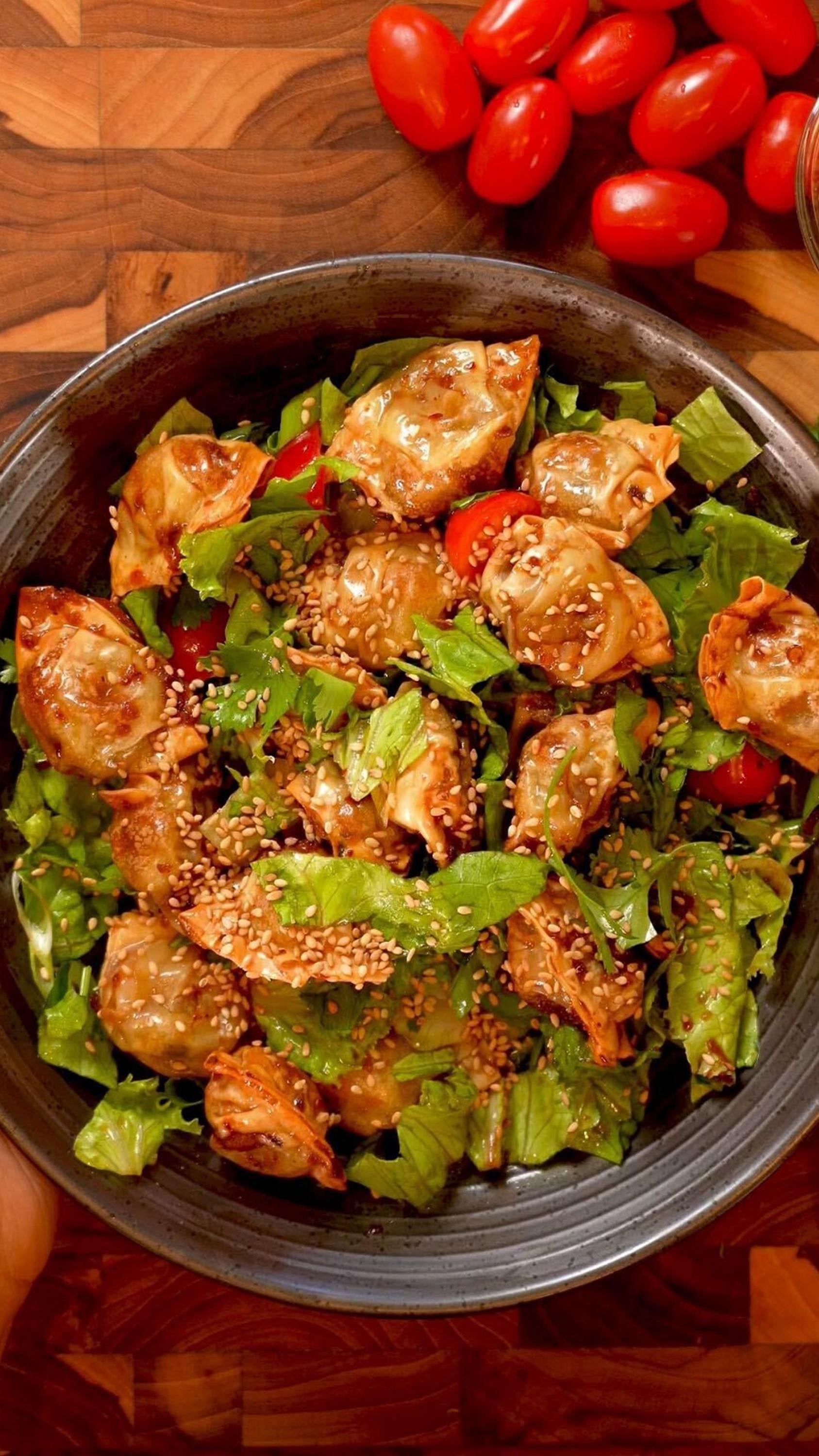 Salad topped with dumplings, sesame seeds, and cherry tomatoes in a bowl on a wooden surface