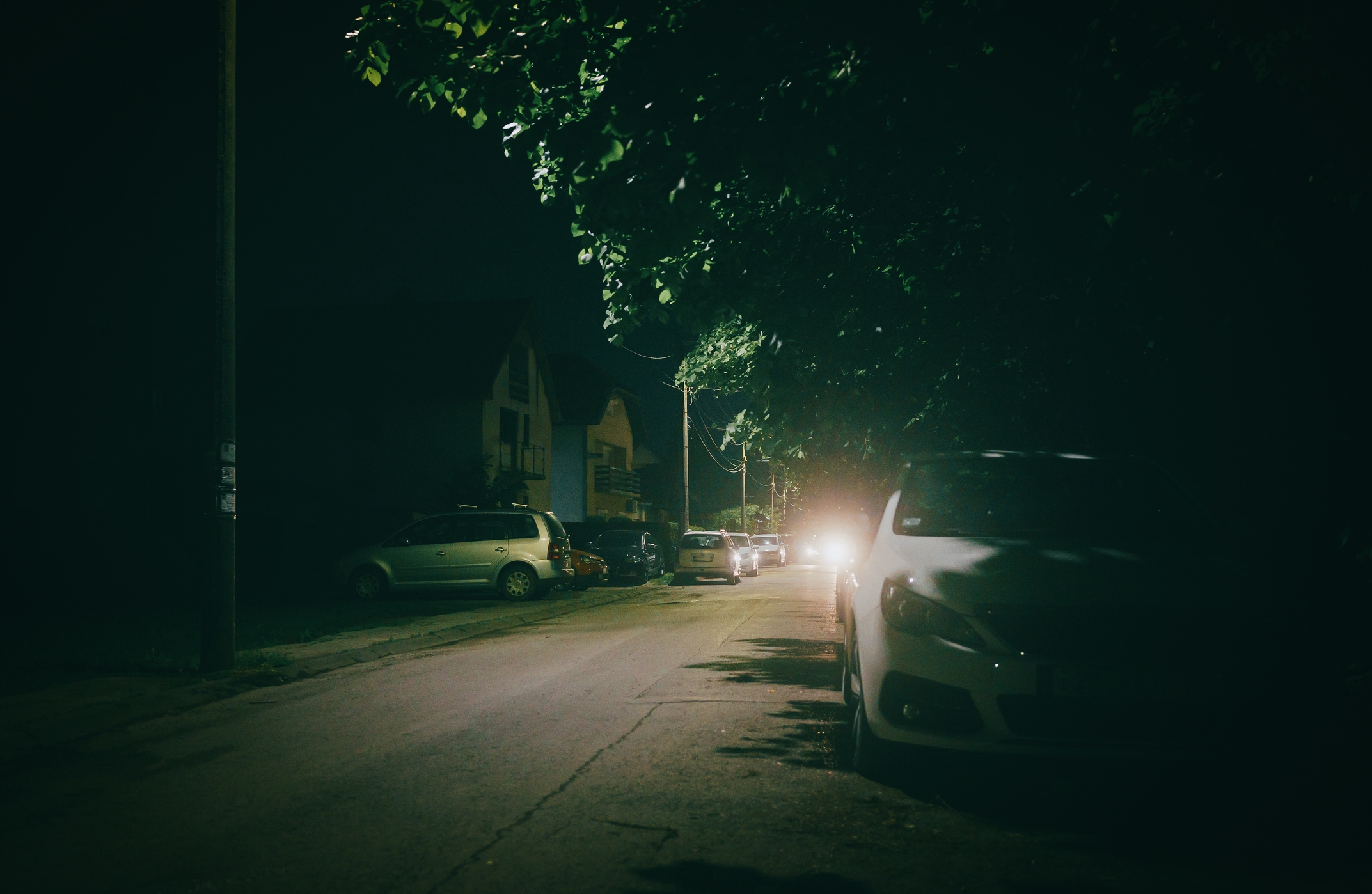 Dimly lit street at night with parked cars and distant headlights casting light on the road