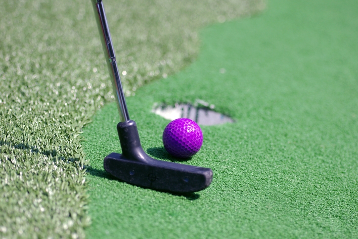 A mini-golf putter positioned to strike a purple golf ball near the hole on a green putting surface
