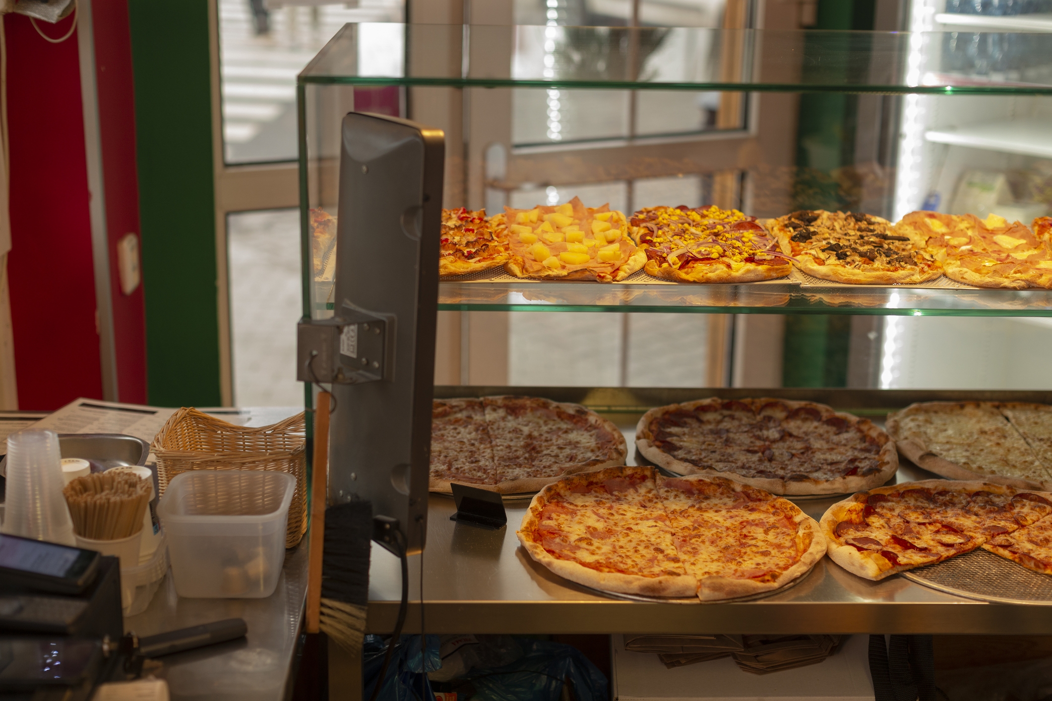 Pizza slices displayed at a counter in a pizzeria, with a variety of toppings visible behind a glass barrier