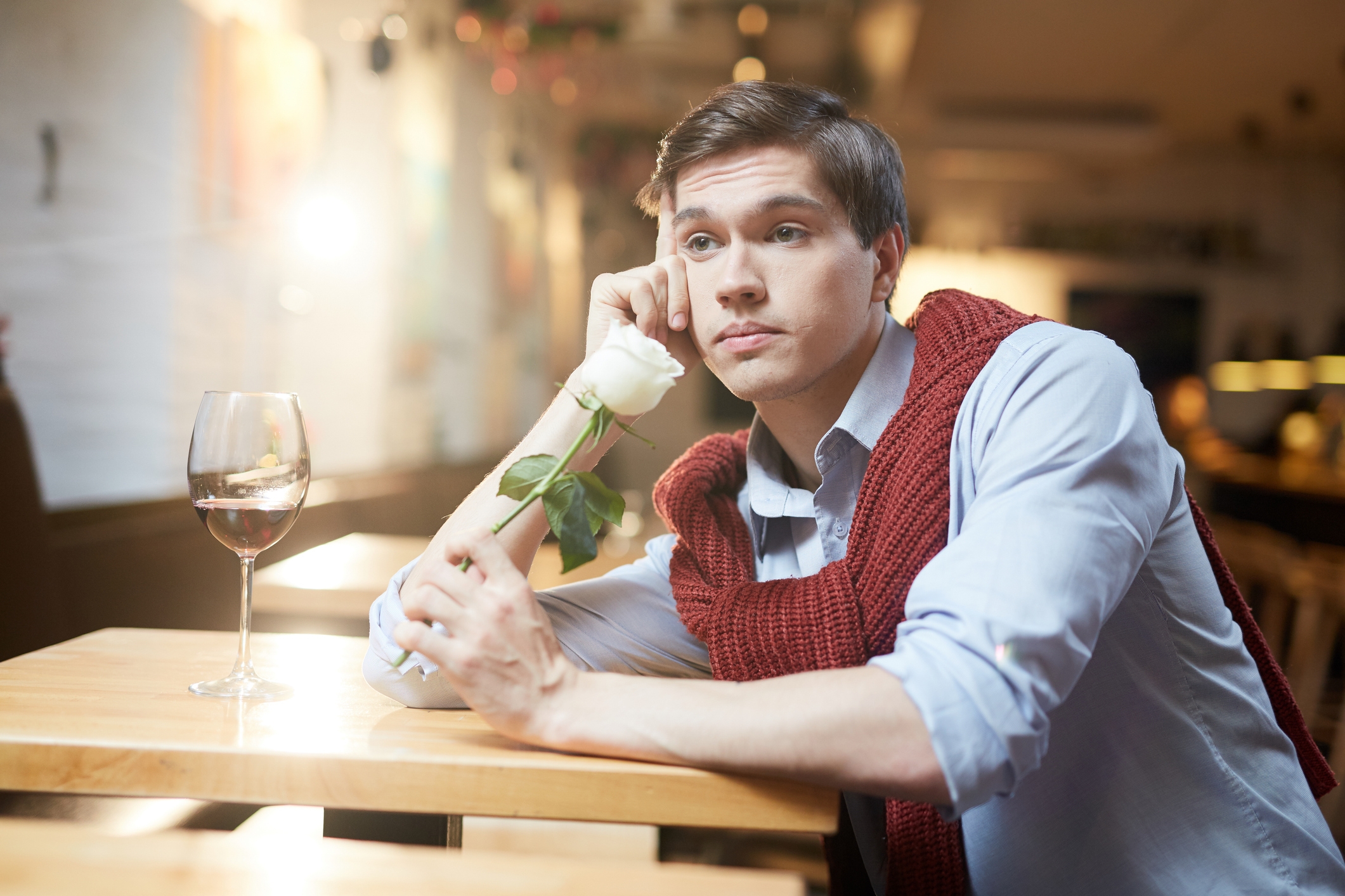Person sitting at a table, holding a white rose and gazing thoughtfully into the distance, with a glass of wine nearby
