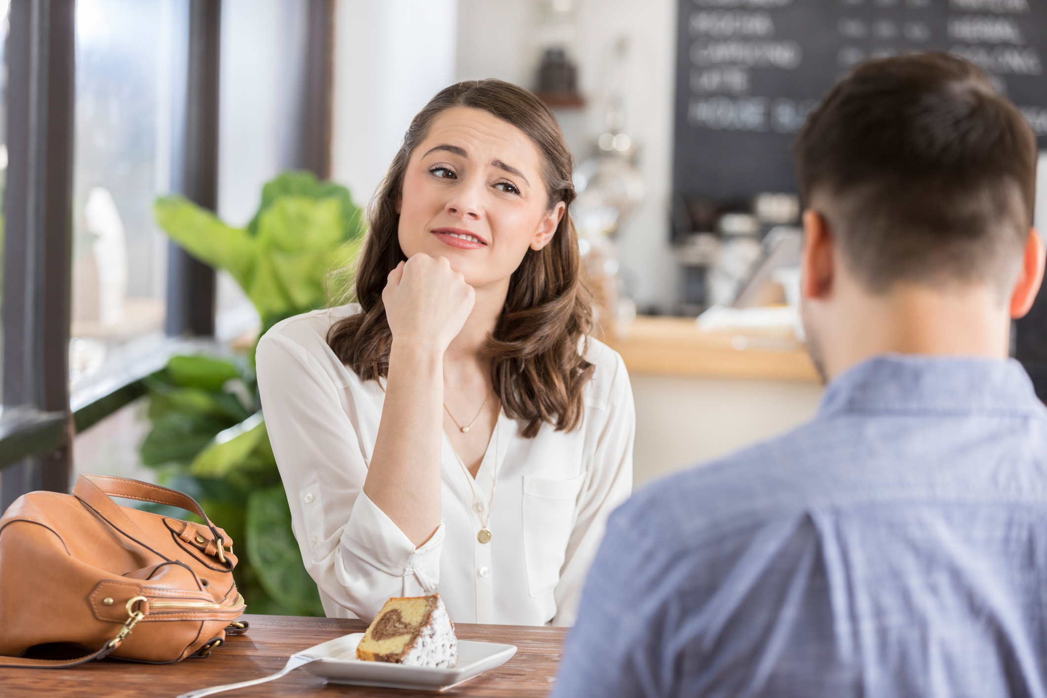 Woman in a cafe looks uncertain during a conversation with a man. A slice of cake and a purse are on the table